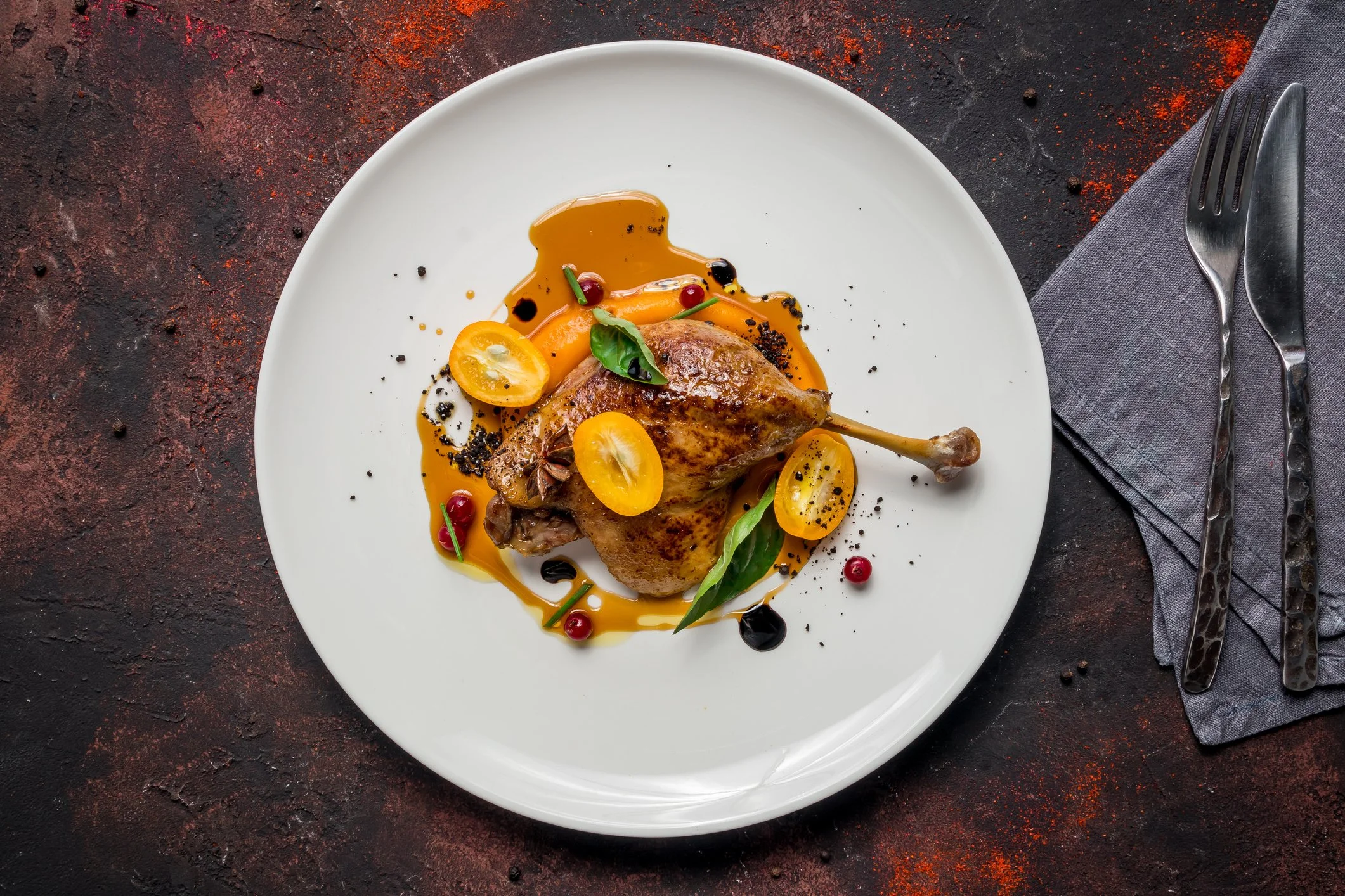 A plated dish featuring a roasted duck leg with orange sauce, garnished with orange slices, green leaves, and berries, on a white plate with silverware on a dark textured surface.