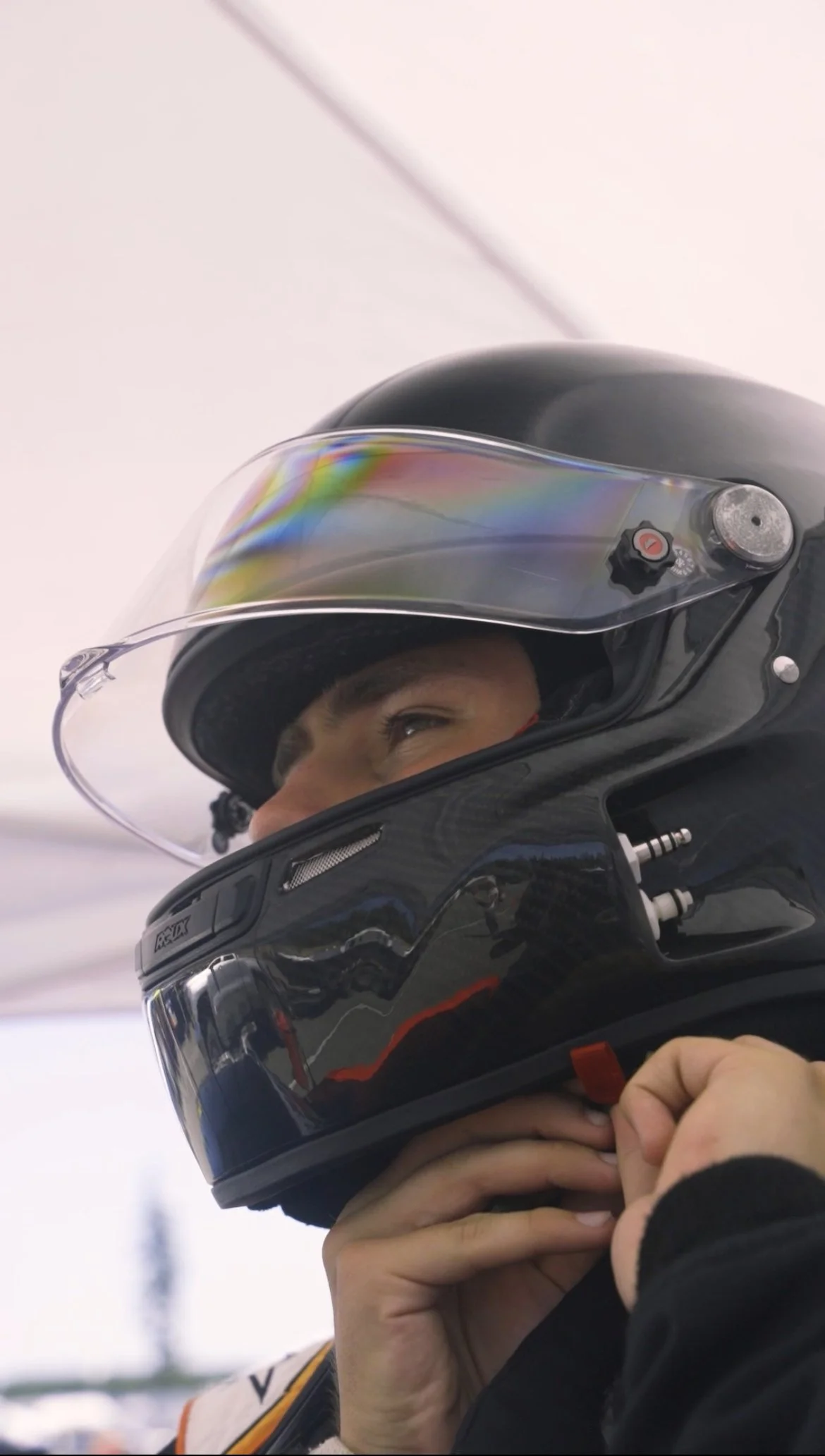 A person wearing a black motorcycle helmet with a clear visor, adjusting or holding the helmet strap with their hands.