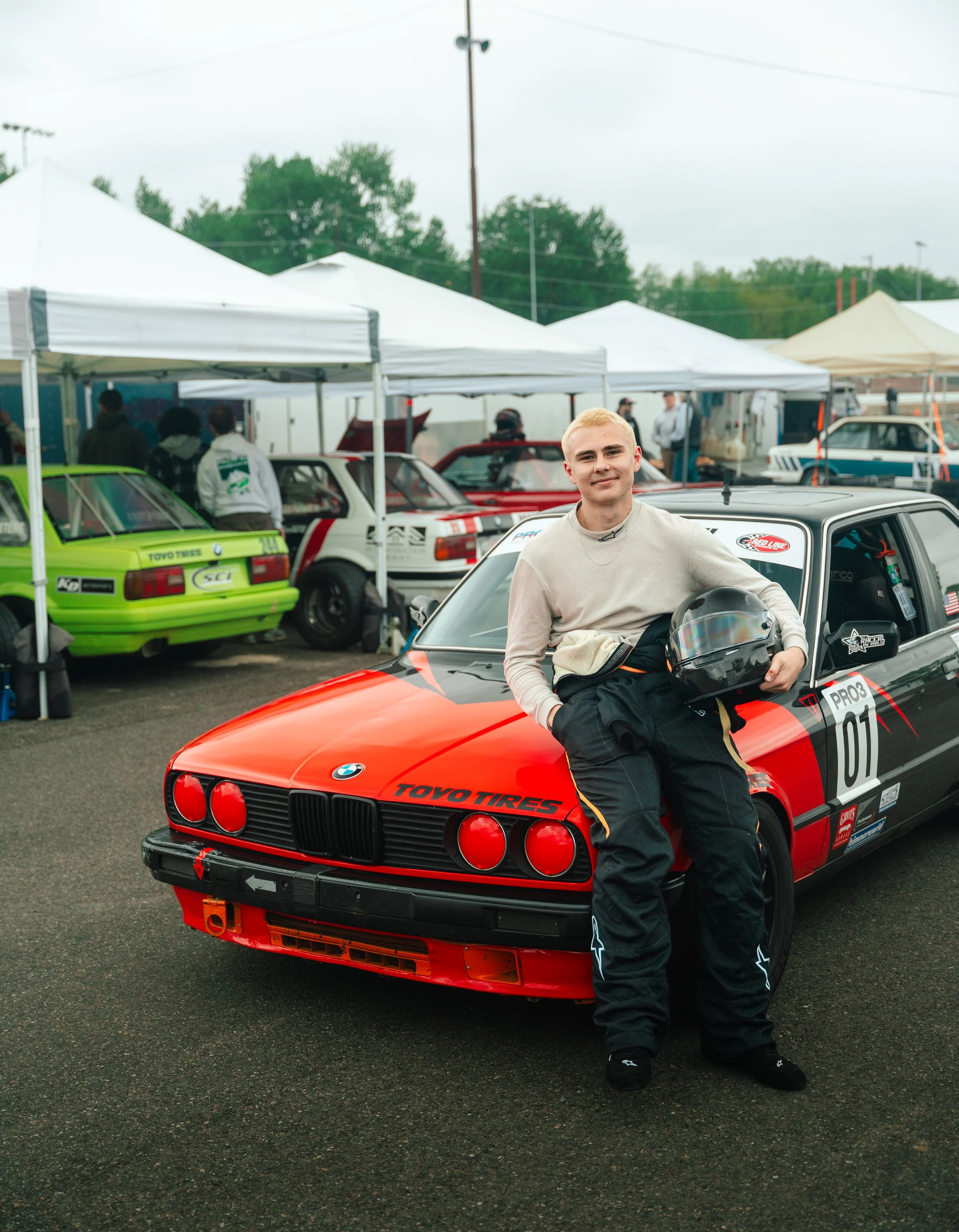 A young man in racing attire sitting on the hood of a red BMW race car at a racing event, holding a black helmet and smiling.