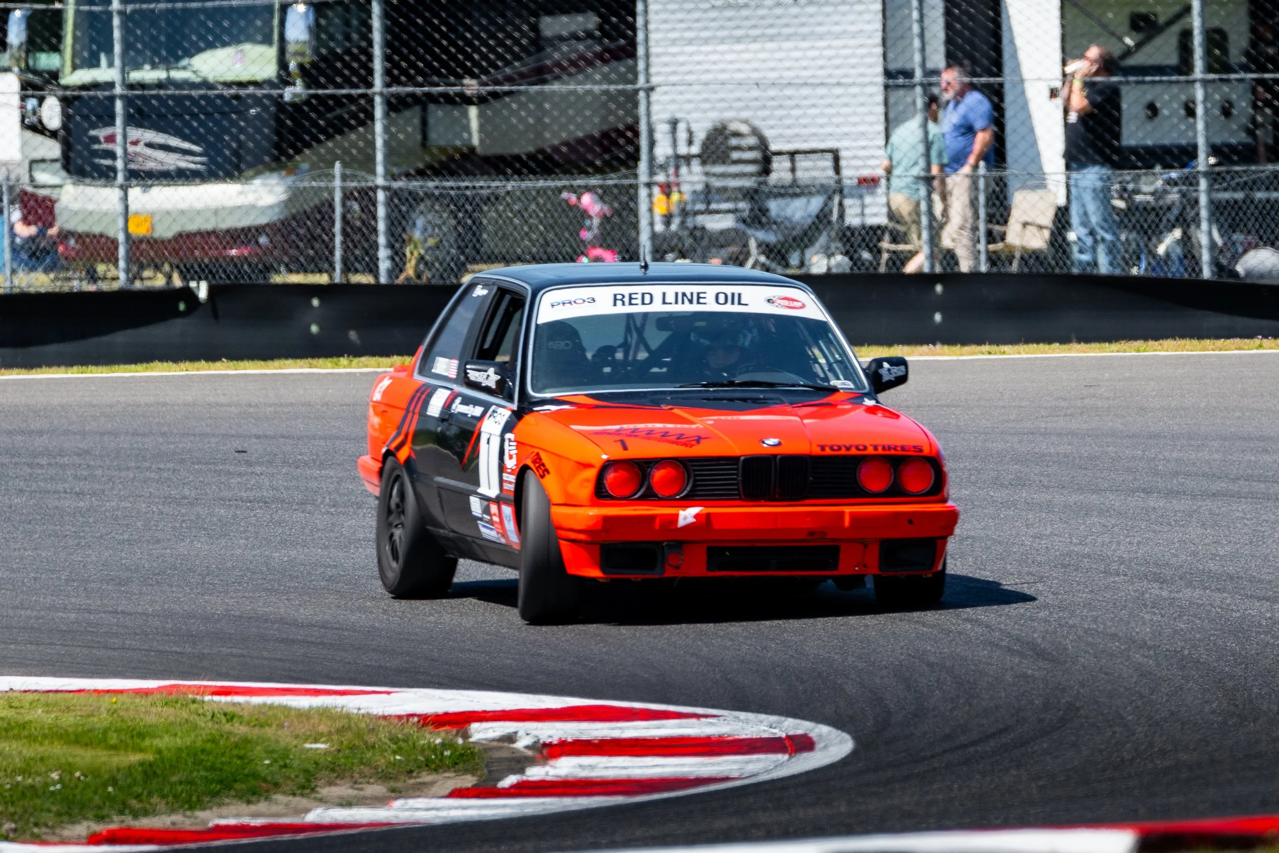 A vintage BMW race car painted orange and black on a race track, with people and tents in the background.