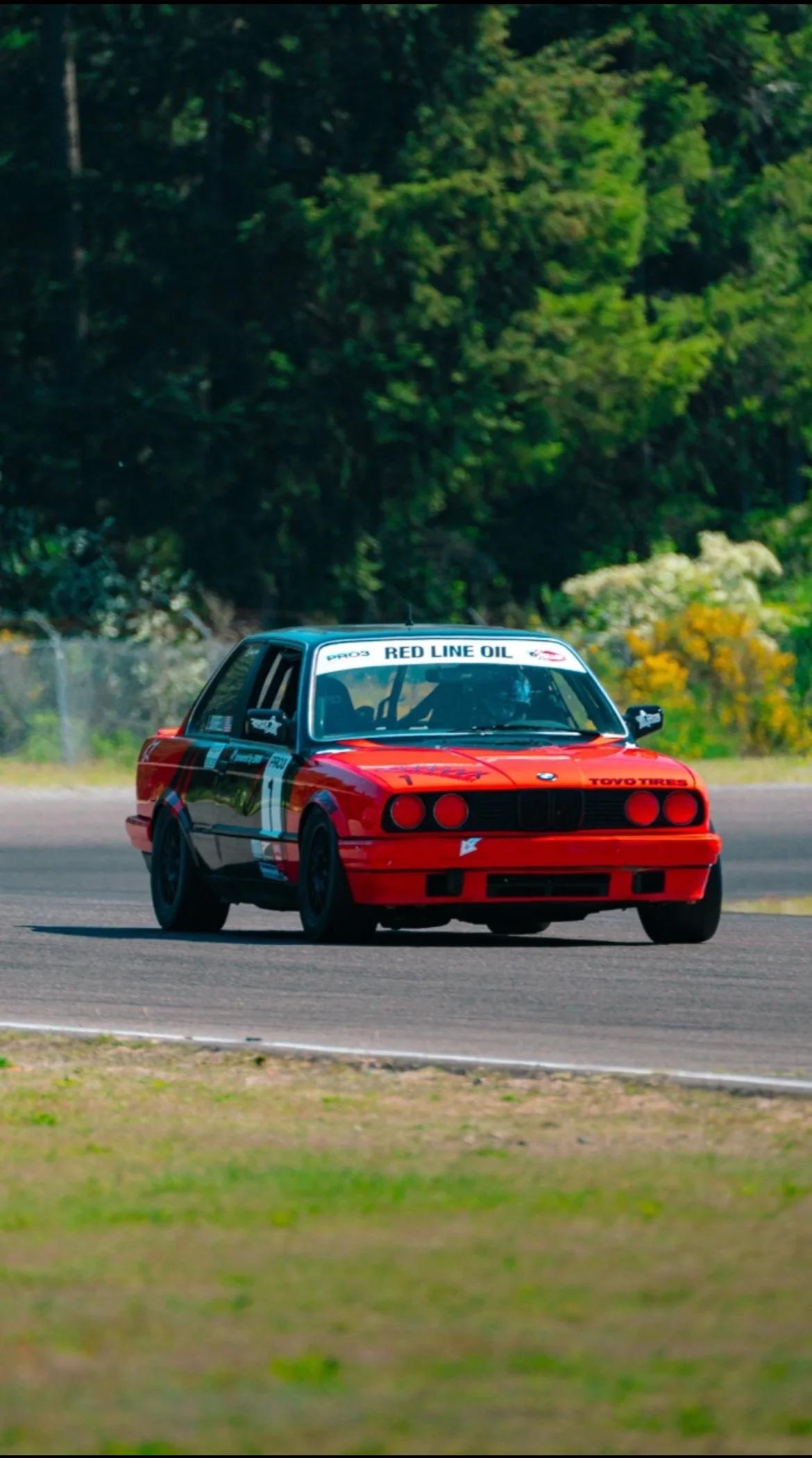 A red race car on a race track during daytime, with a black spoiler and racing decals, surrounded by green trees and bushes.
