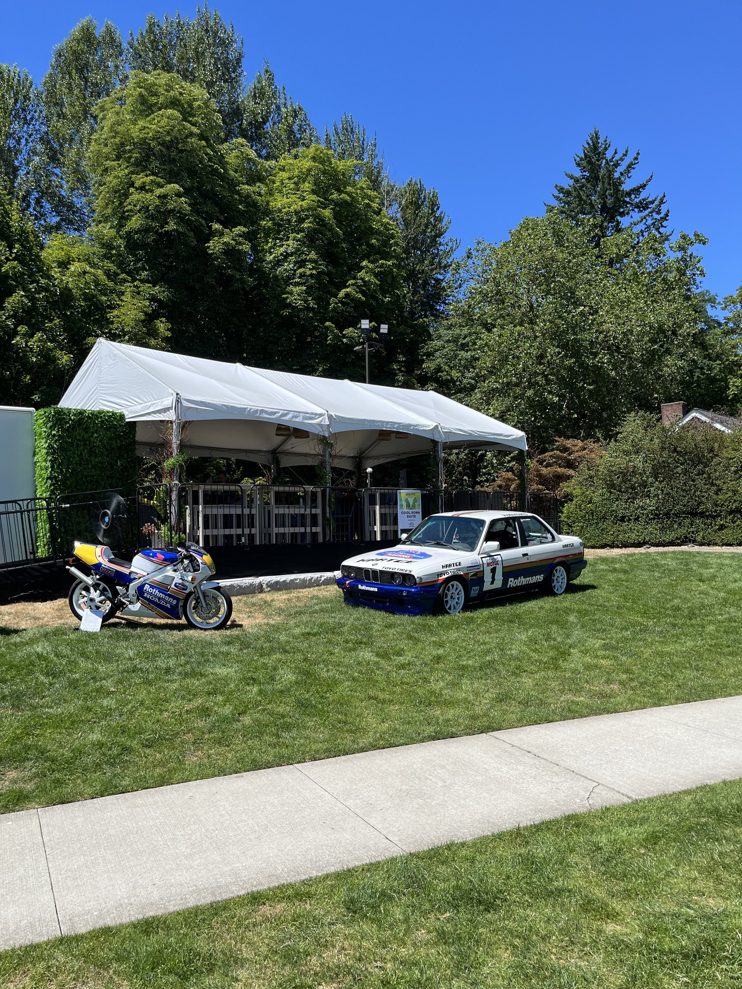 Display of vintage motorsport cars and a motorcycle outside under a white tent with trees and a blue sky in the background.