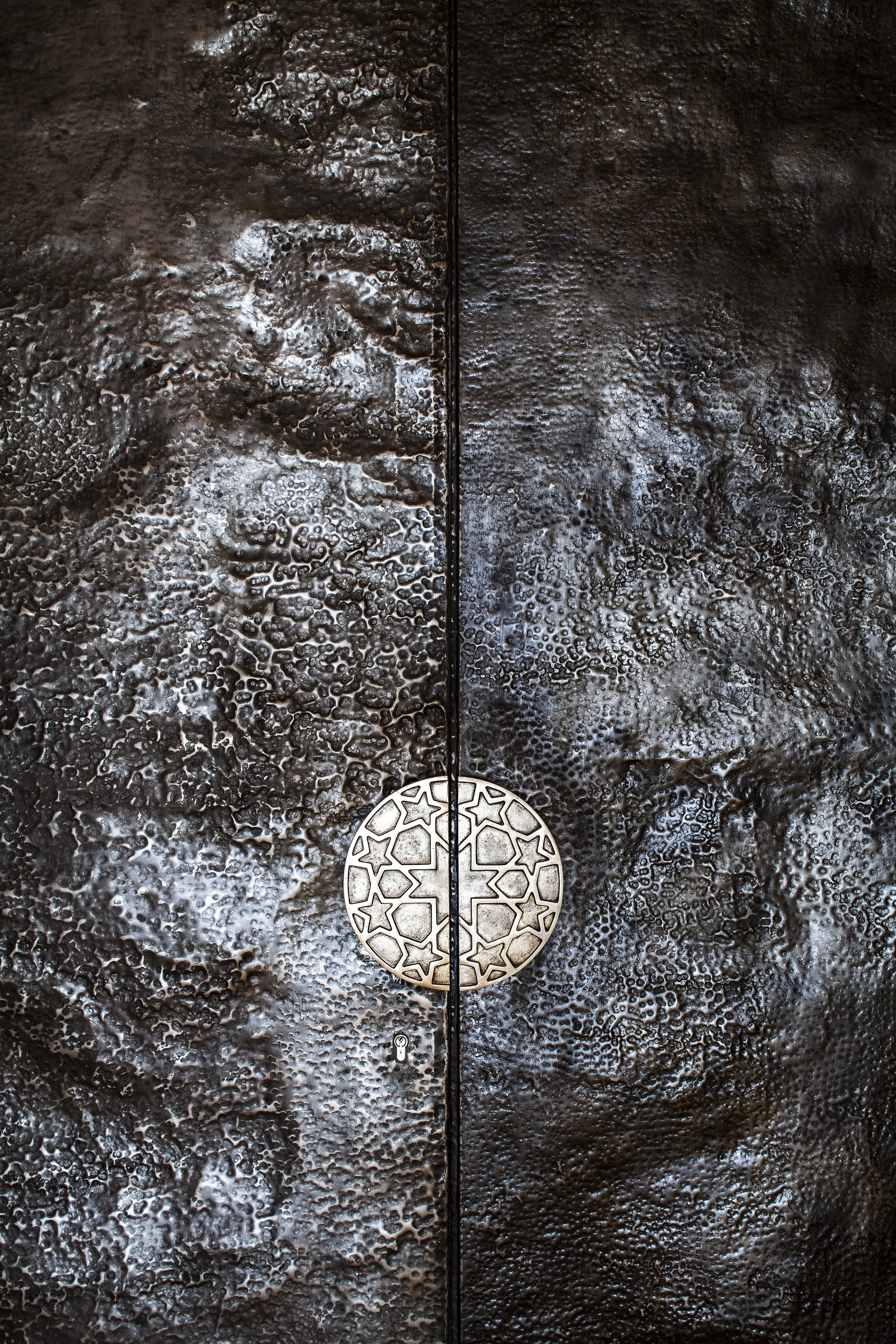 Close-up of a metal door with a textured, hammered metal surface texture, and a round silver handle with geometrics.