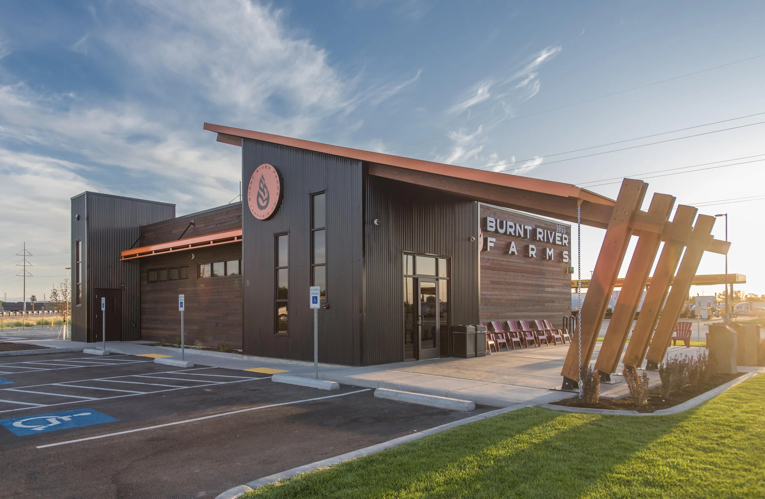 Exterior view of Burnt River Farms building with a modern design, black siding, and pink accents, featuring a large wooden sculpture in the foreground, a parking lot with designated handicapped spots, and a blue sky with scattered clouds.