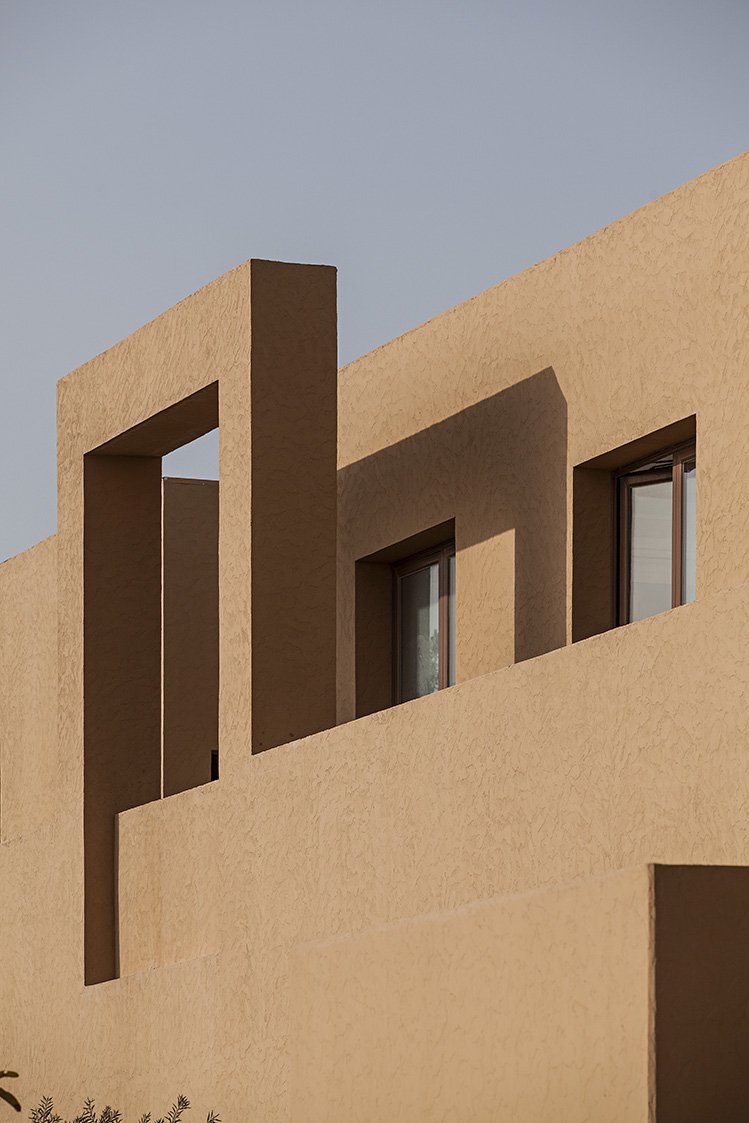 Close-up of a modern beige stucco building with rectangular windows and geometric architectural design, shadows cast on the wall, blue sky in the background.