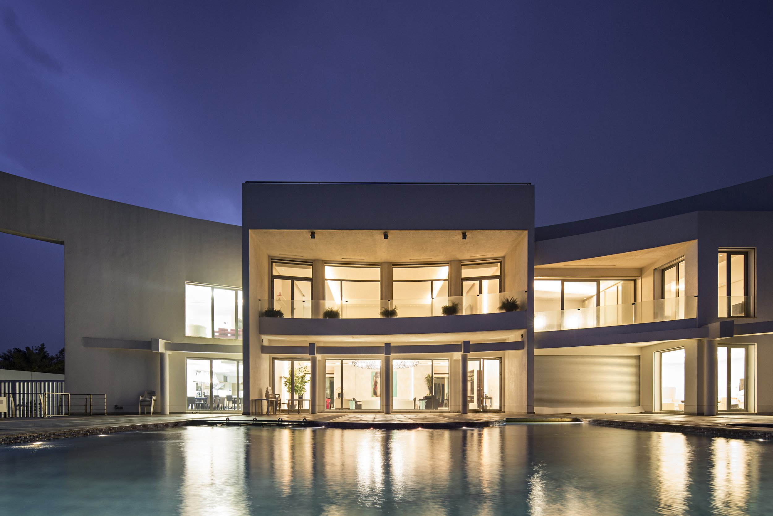 Modern two-story house with illuminated interior, large glass windows and balcony, reflecting in a swimming pool at night.