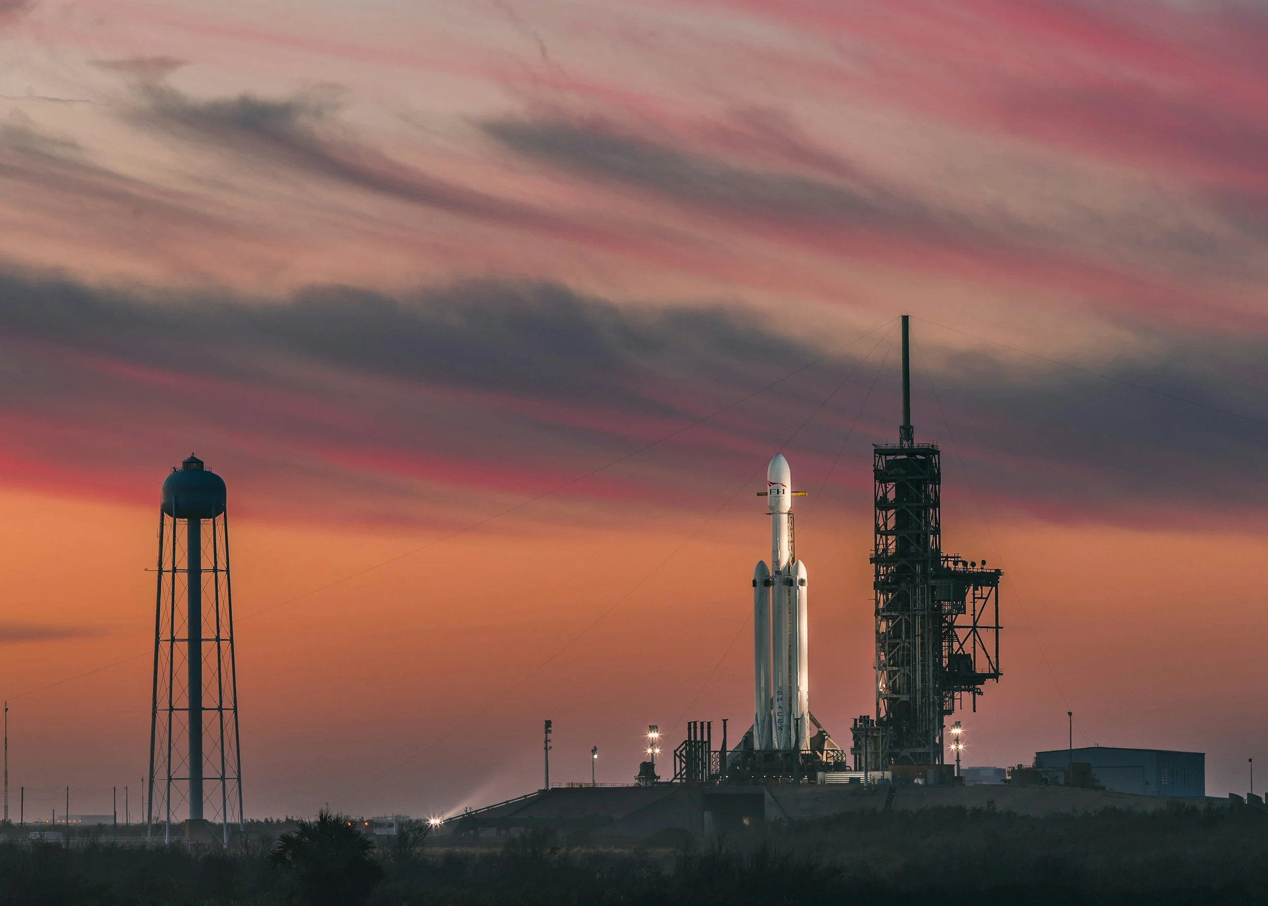A rocket on a launch pad at sunset, with a colorful sky and surrounding structures including a water tower.