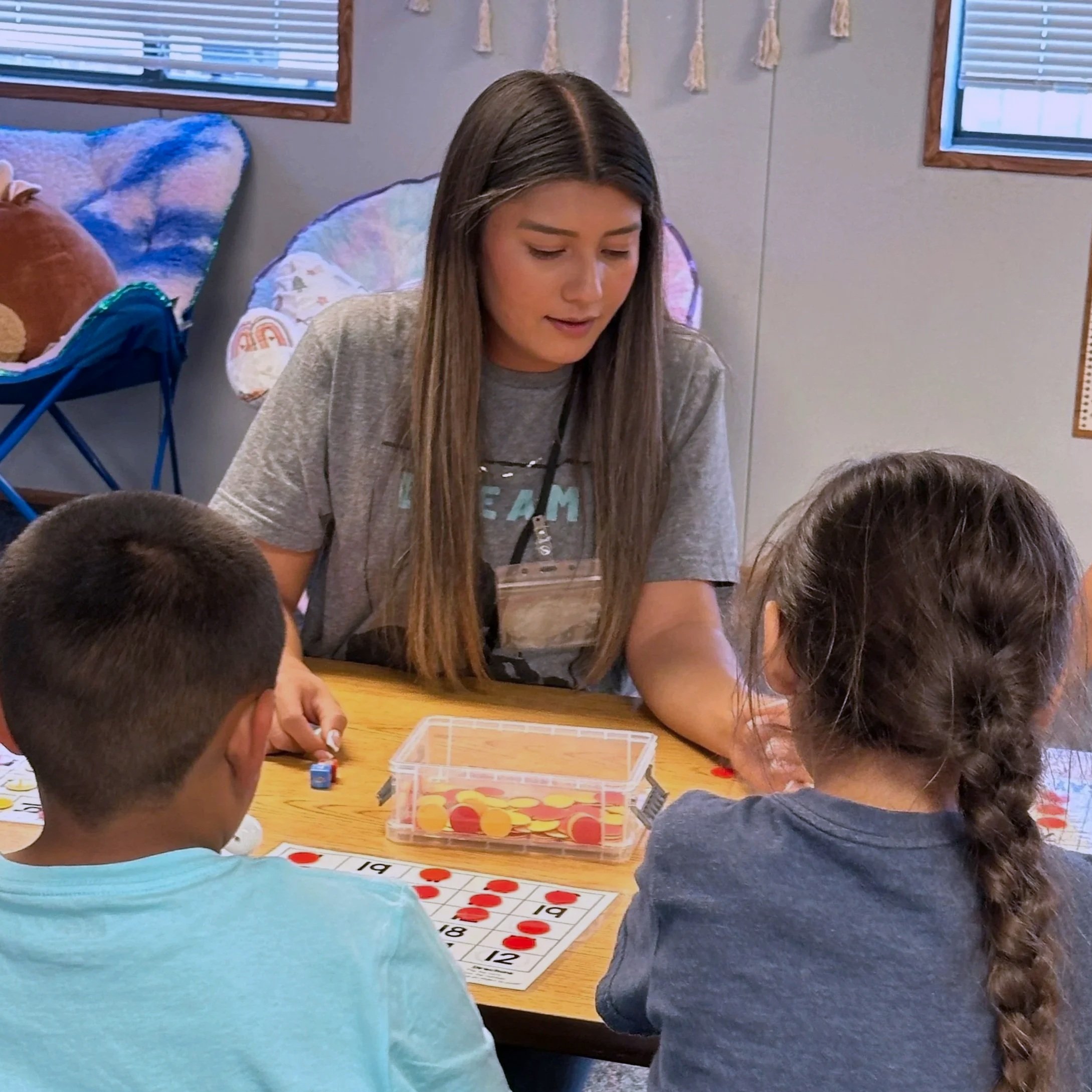 A group of children and a young woman sitting around a table playing a game of Bingo in a classroom or recreational room. The table has a bingo card with red markers, and a transparent plastic container holding the markers.