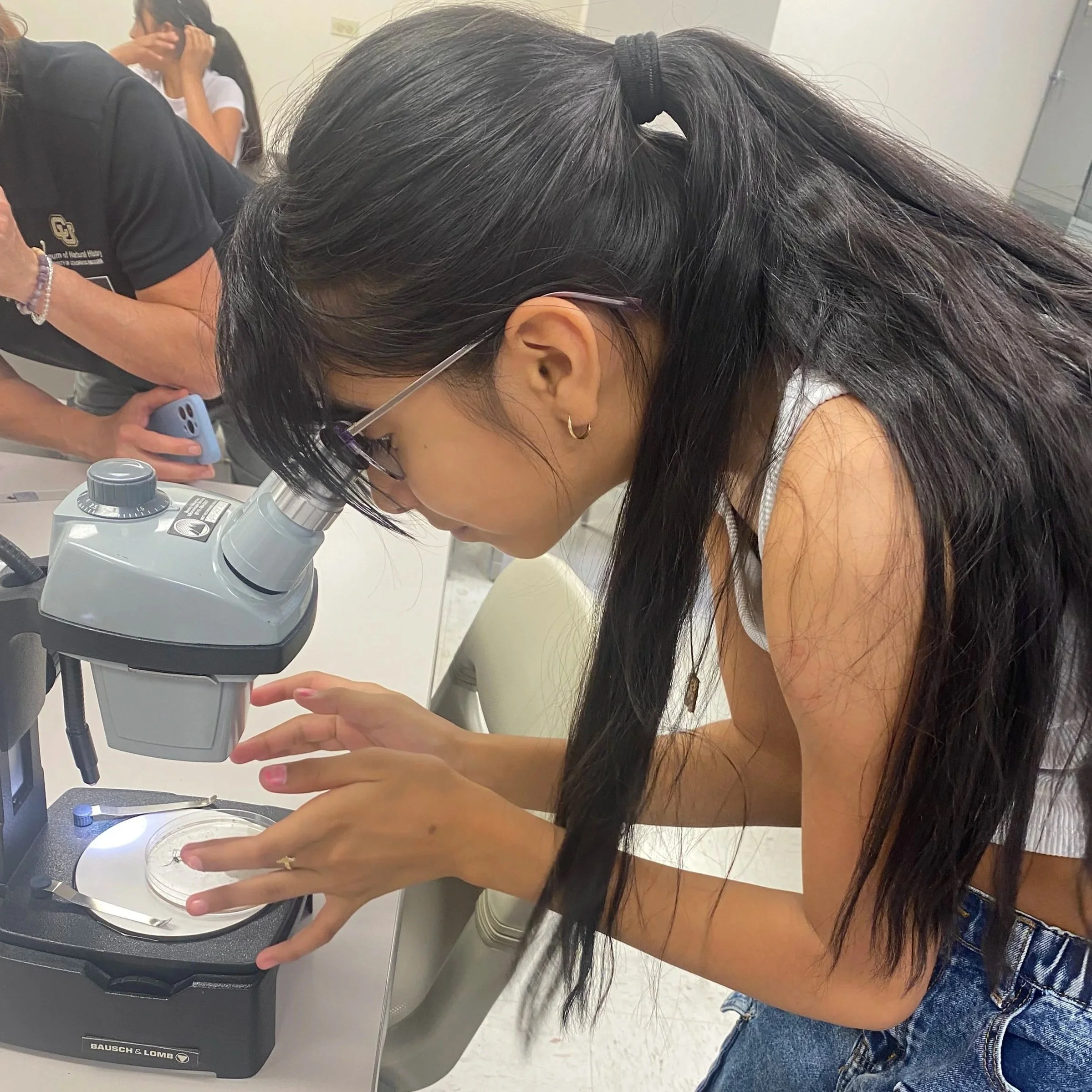 A woman with long dark hair wearing glasses and a white tank top is looking into a microscope with her right hand holding her hair back. The microscope is a Bausch & Lomb brand, and there is a person in the background wearing a black t-shirt and holding a smartphone.