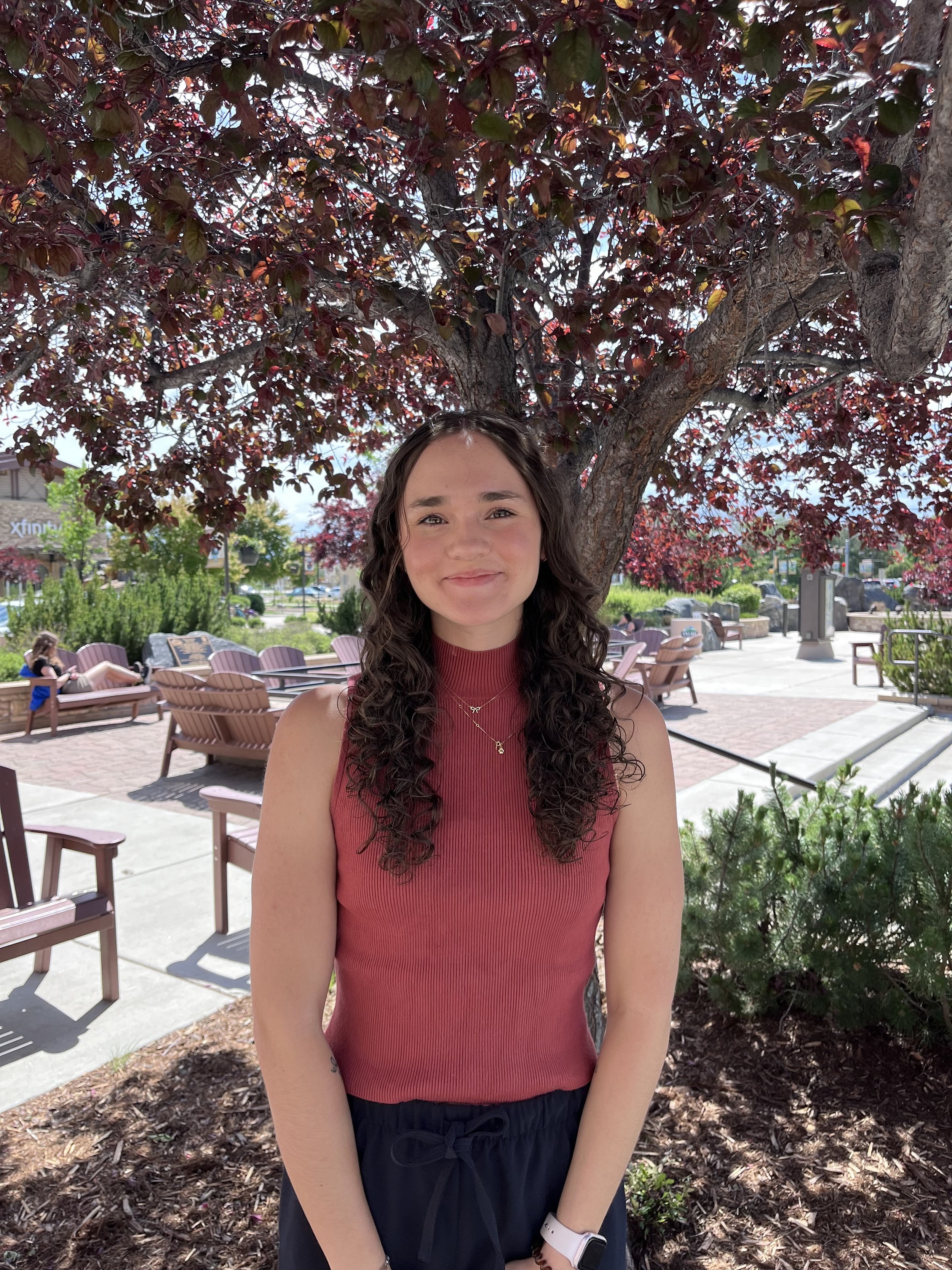 A young woman with curly brown hair smiling outdoors near a tree with dark red leaves. She is wearing a sleeveless red top, dark pants, and a smartwatch on her left wrist.