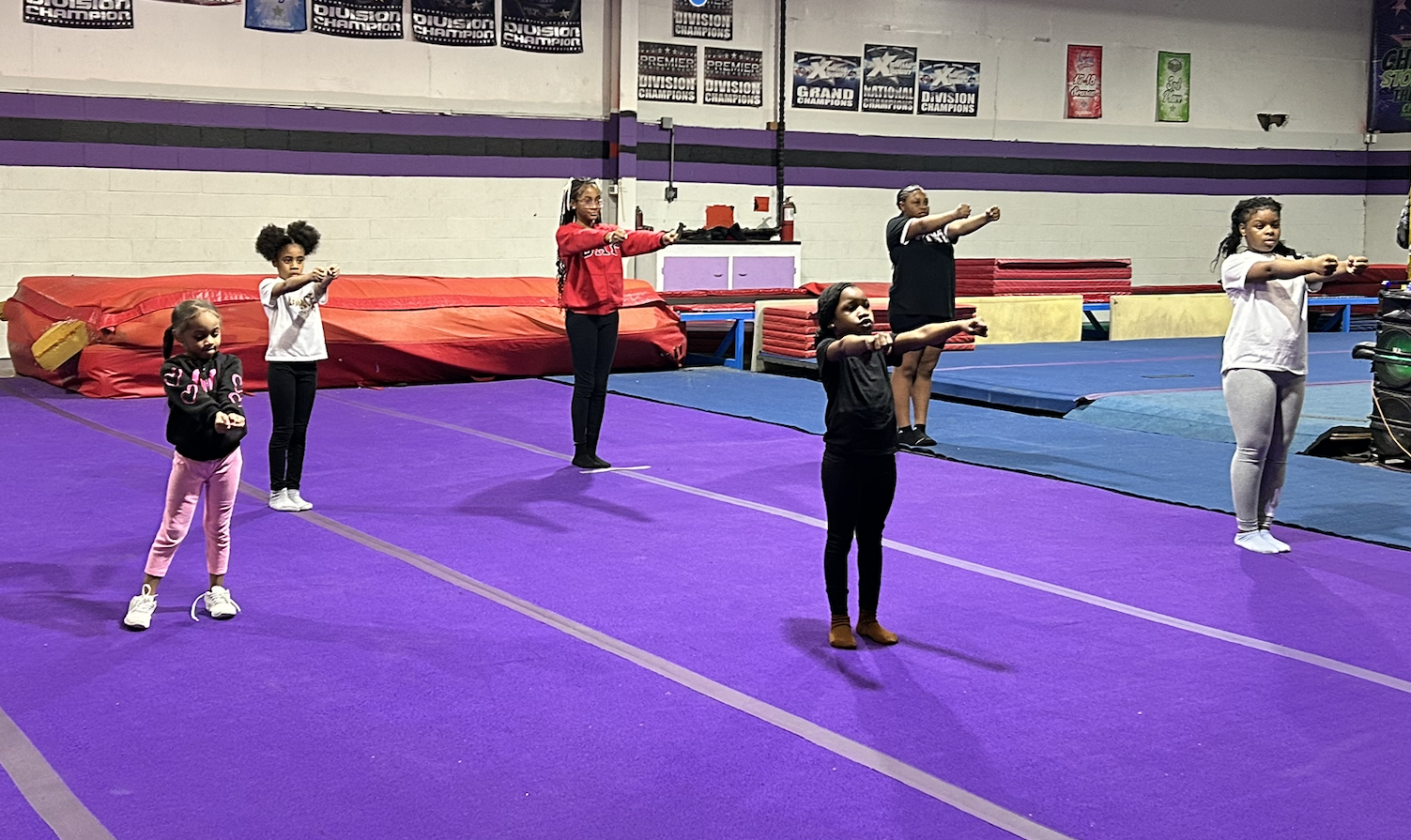 Six young girls practicing gymnastics on purple mats in a gymnasium with posters on the wall.