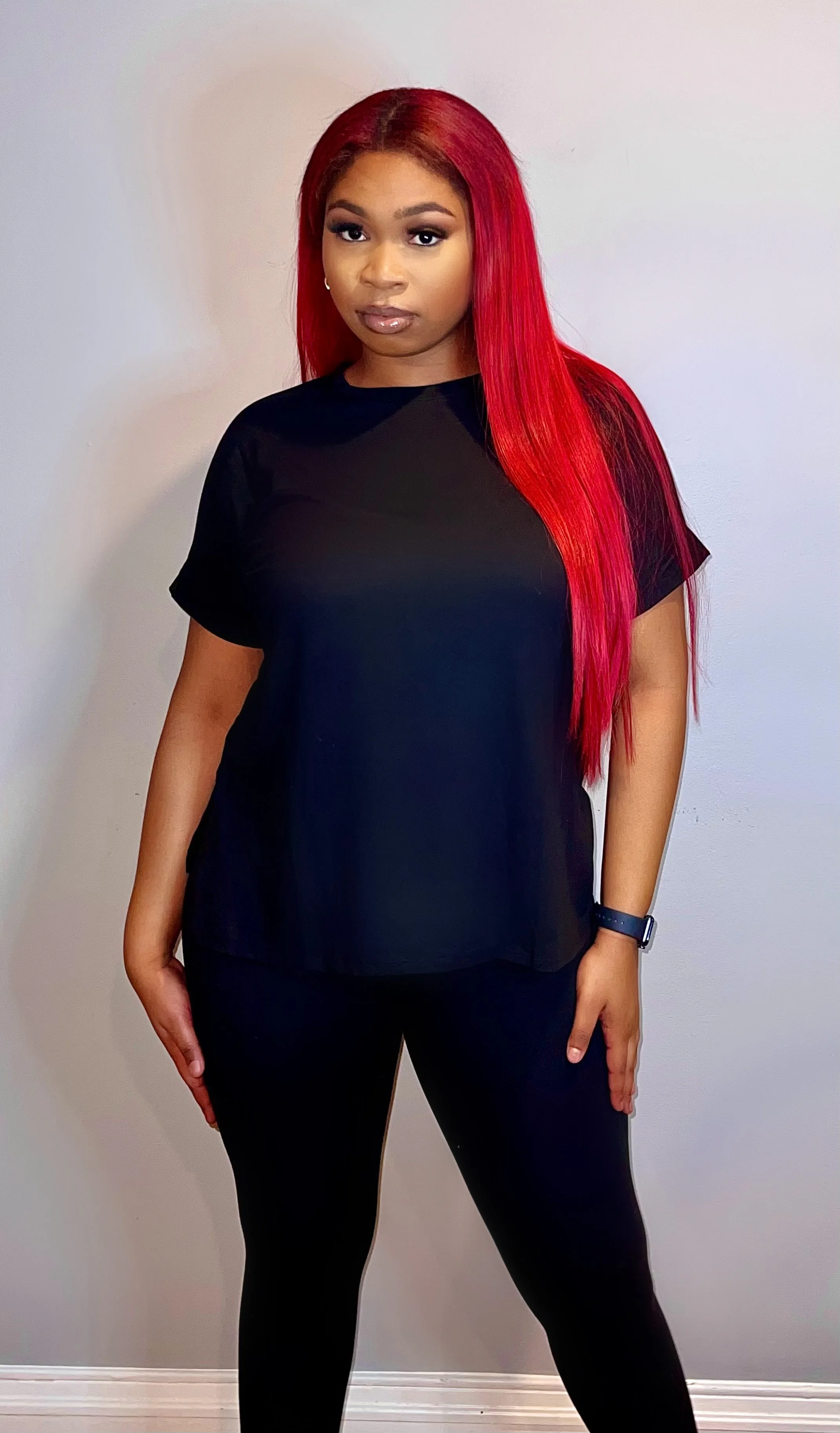 A woman with long red hair wearing a black t-shirt, black leggings, and a wristwatch, standing against a plain white wall.