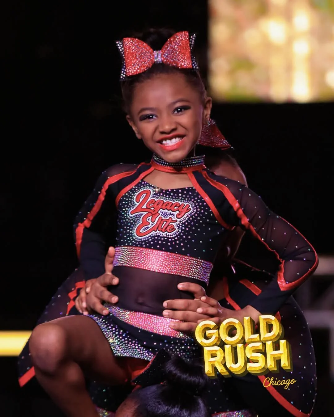 Young girl in a sparkly cheerleading uniform with 'Legacy Elite' on the front, smiling confidently, with a large red bow in her hair, performing a cheerleading pose at a competition.