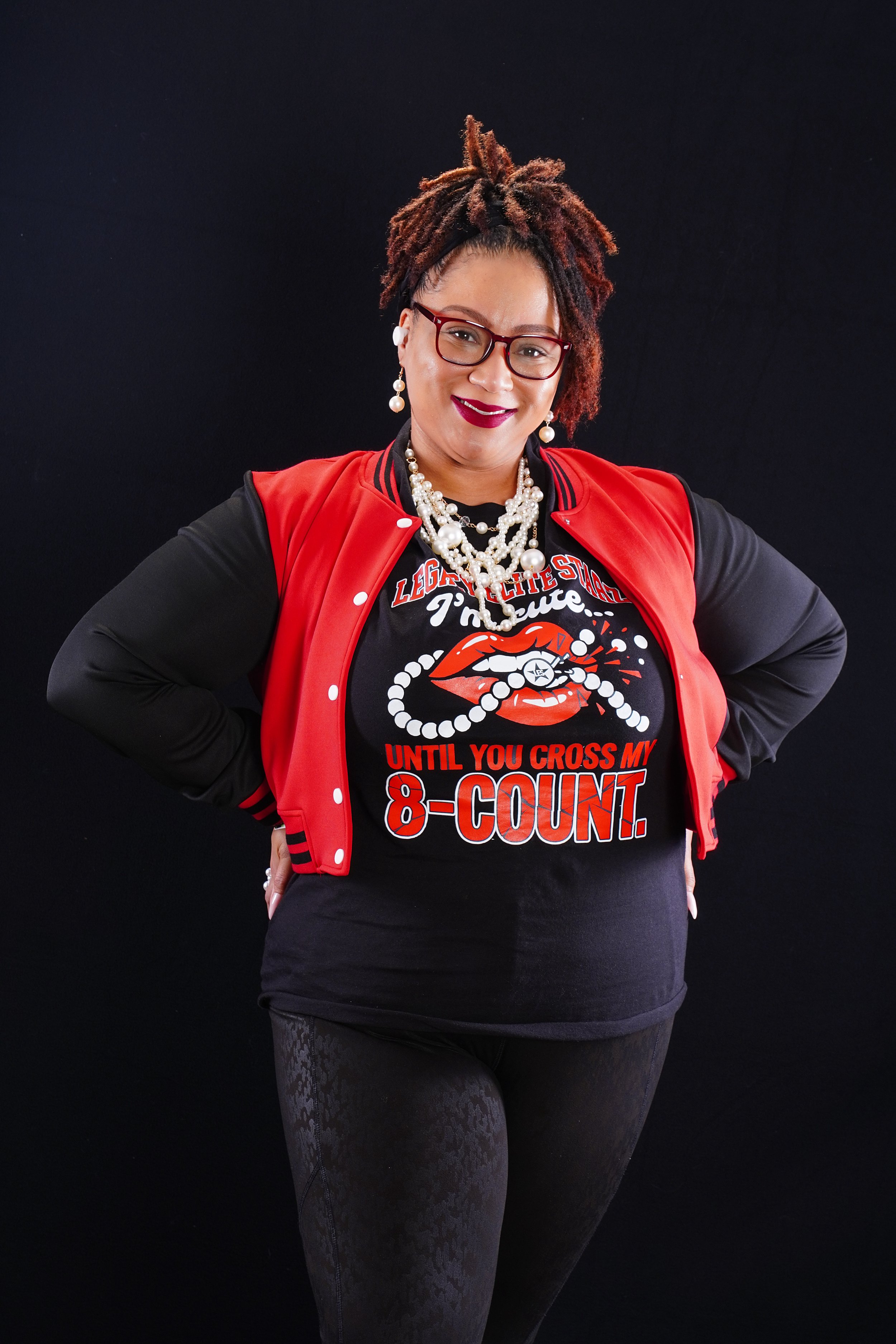 A woman wearing glasses, a red varsity jacket, and layered pearl jewelry, standing against a black background. She has dreadlocks and is smiling while wearing a black t-shirt with a graphic and slogan.