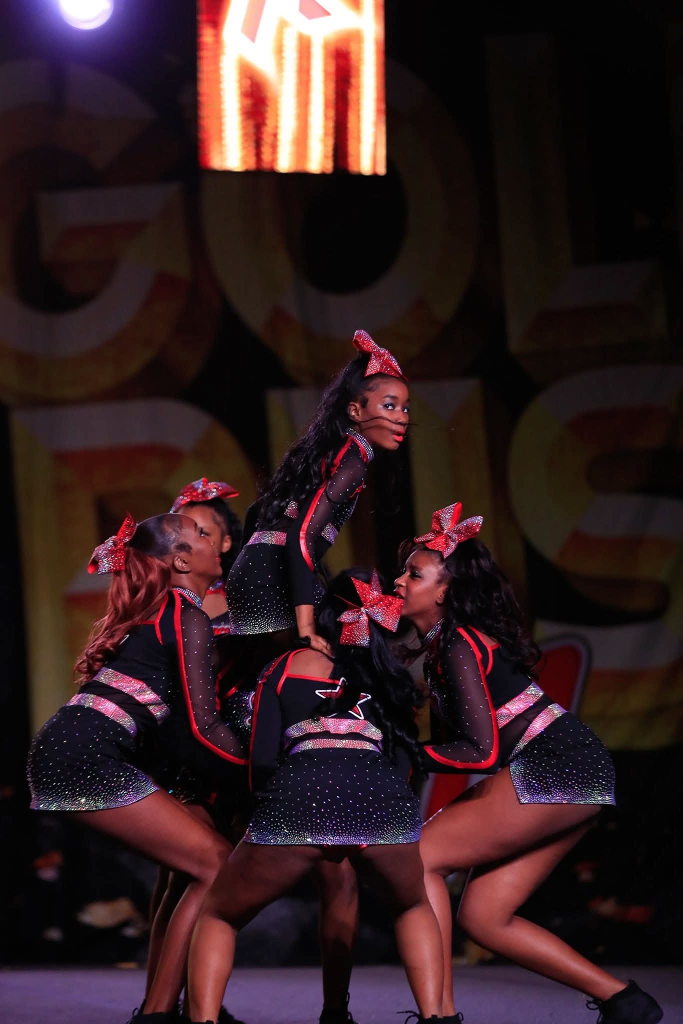 A group of young female cheerleaders performing a stunt on stage. They are dressed in black and red uniforms with sparkly embellishments, and red bows in their hair. The cheerleader at the center is lifted by her teammates.