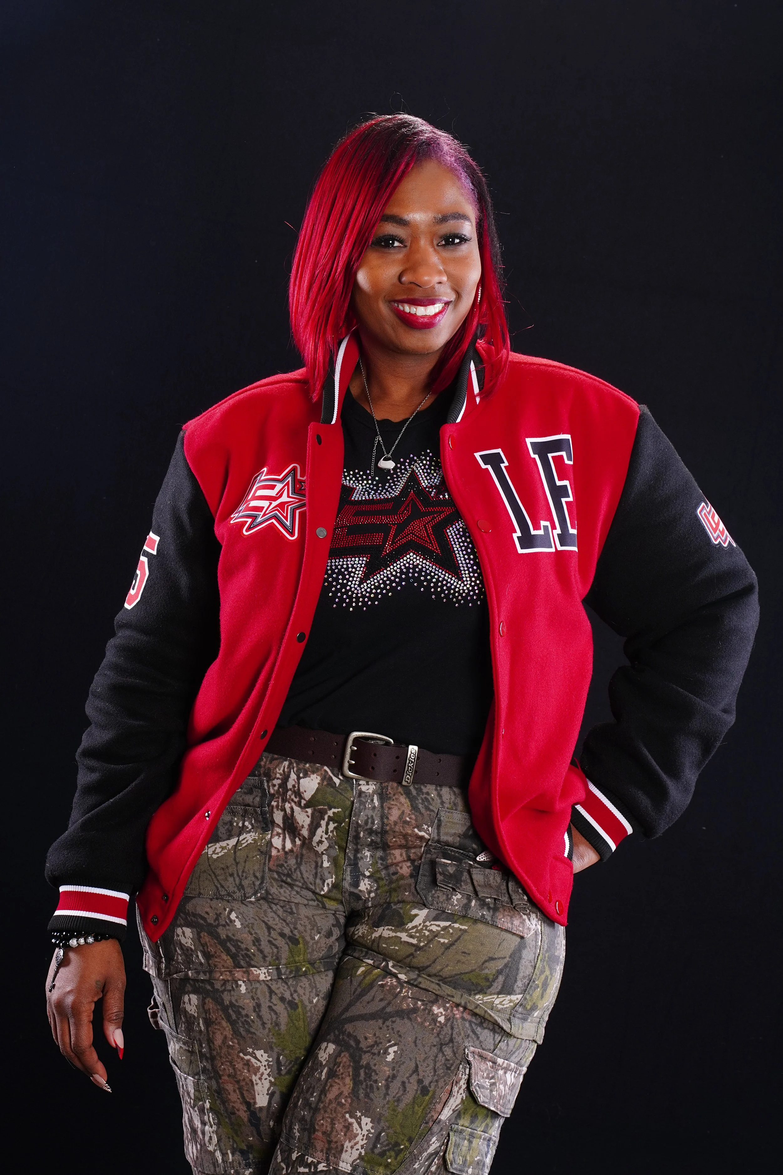 Woman with pink and purple hair in a red and black jacket with patches and design, Camouflage cargo pants, smiling, posing against black background.