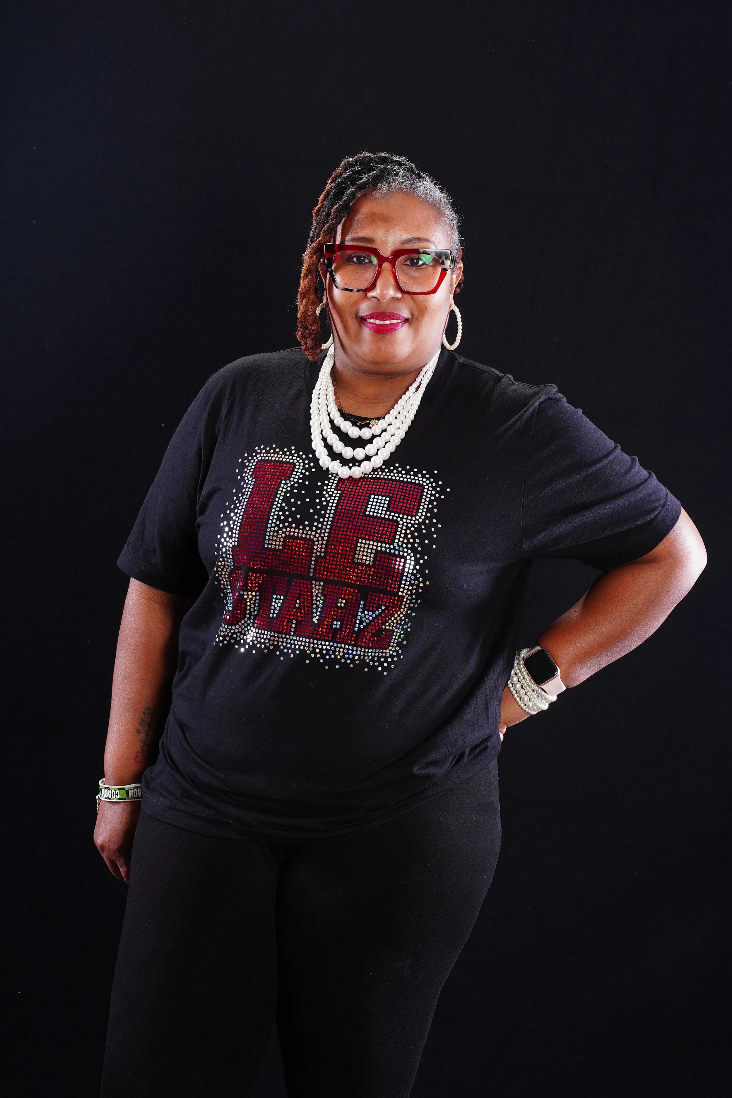 A woman with dark skin, glasses, and dreadlocks posing against a black background. She is wearing a black T-shirt with a sparkling design and the words "LIFE IS A BAZZ" on it, along with multiple pearl necklaces, hoop earrings, and a white wristwatch.