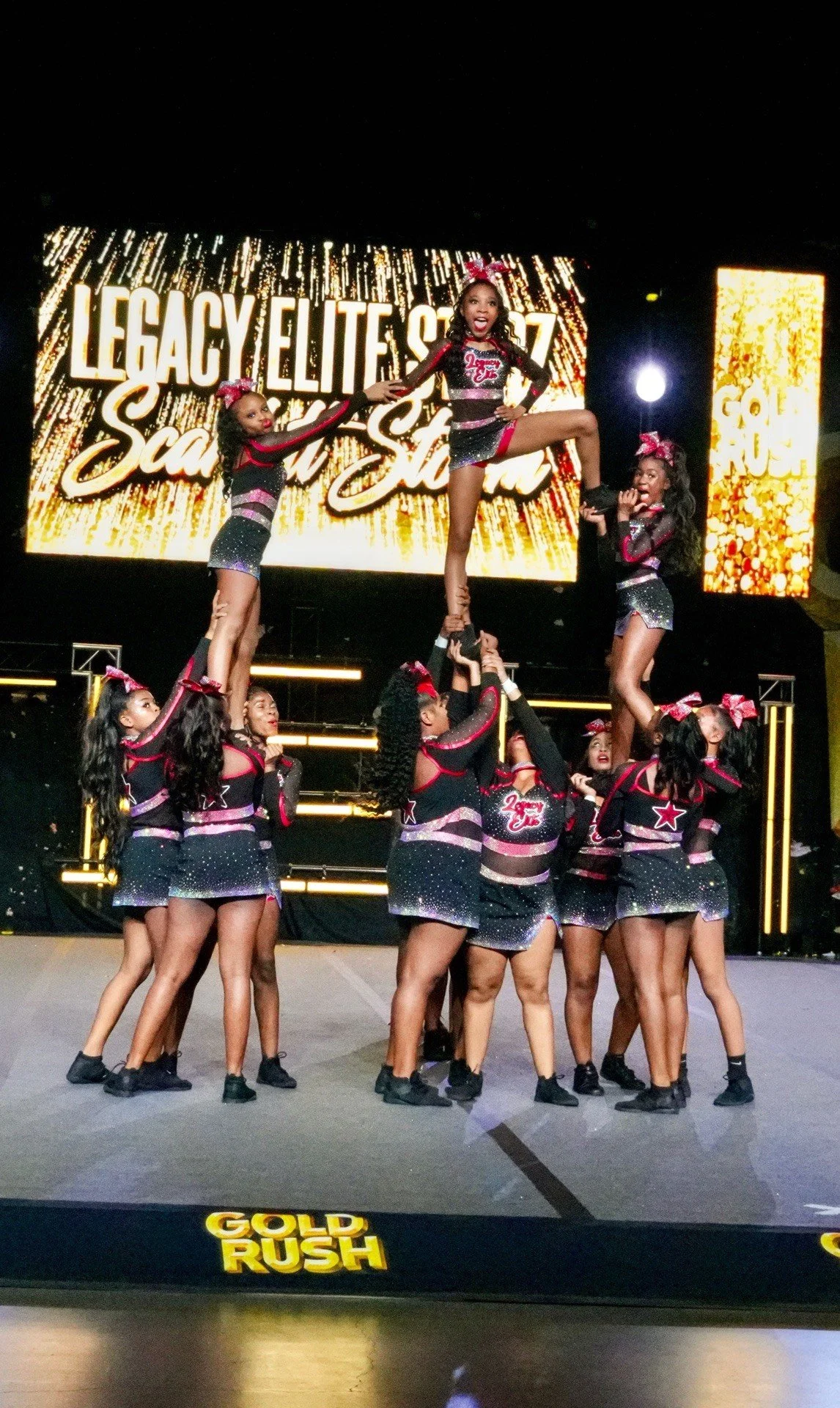 Cheerleaders performing a routine on stage during a competition with a large screen behind them displaying the words "Legacy Elite" and "Star".