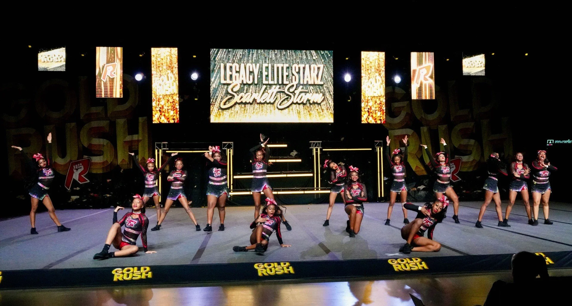 A group of young cheerleaders in black and red uniforms performing a dance routine on stage at a cheerleading competition, with a large screen behind them displaying "Legacy Elite Starz Scarlett Storm."
