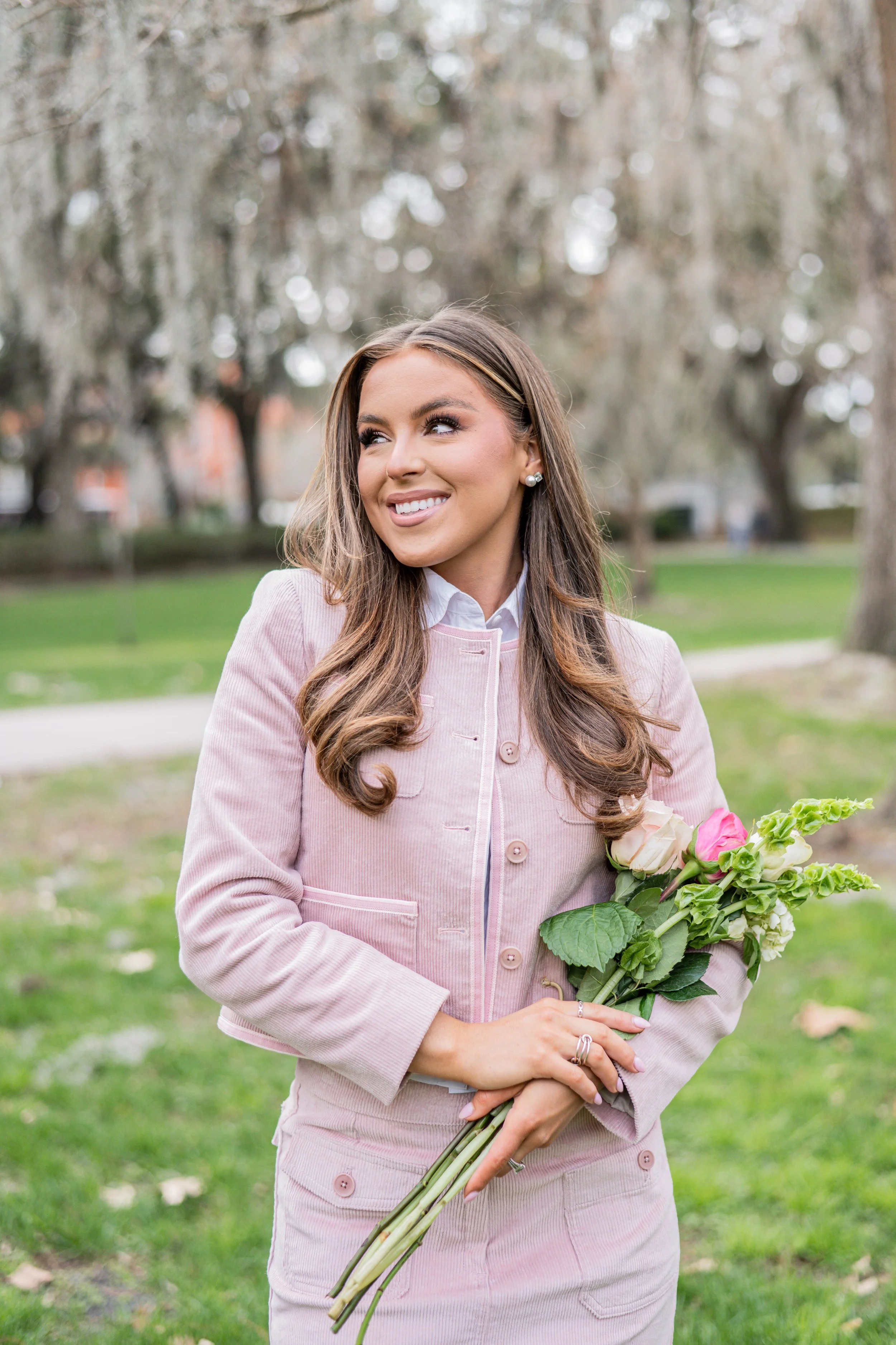 A woman in a pink jacket holding a bouquet of flowers outdoors with trees and grass in the background.