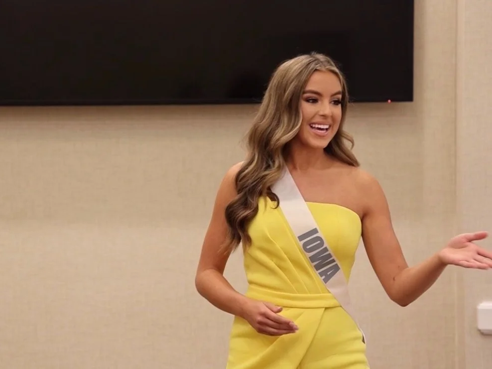 A woman with long, wavy hair wearing a yellow dress with a sash that says 'IOWA' is speaking and gesturing with her hand in an indoor setting.