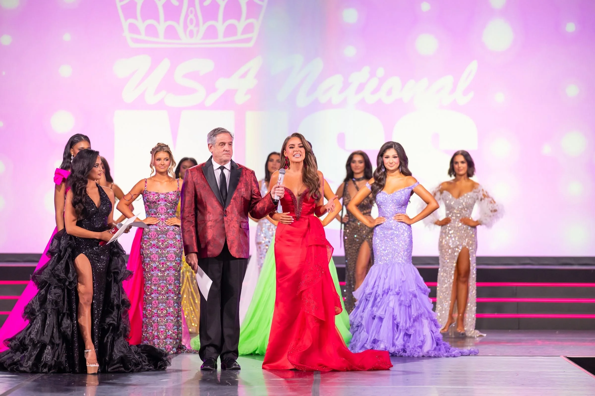 Beauty pageant contestants on stage with a host holding a microphone and a man standing beside her, all dressed in colorful evening gowns and suits.