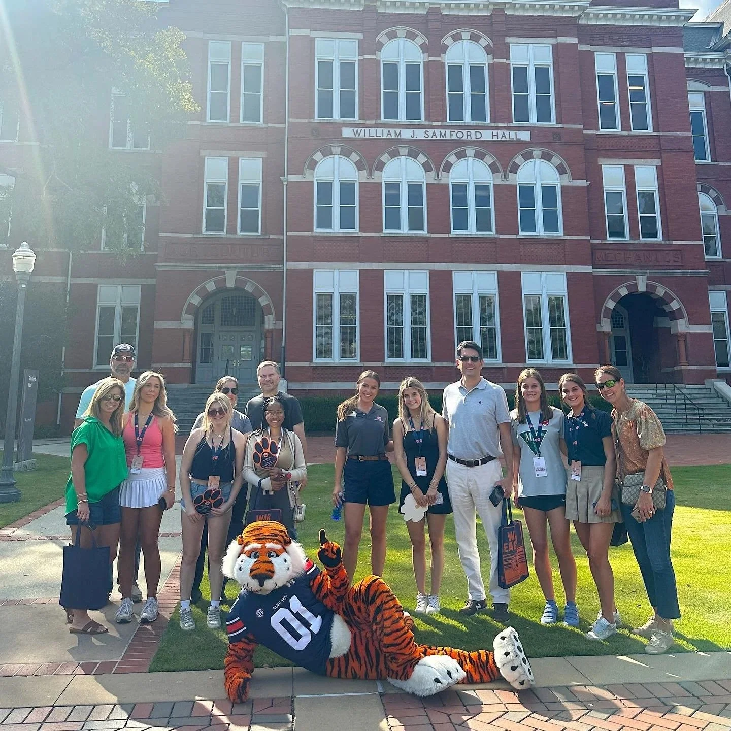 Group of people standing in front of William J. Samford Hall at Auburn University, with a person in a tiger mascot costume lying on the ground in front.