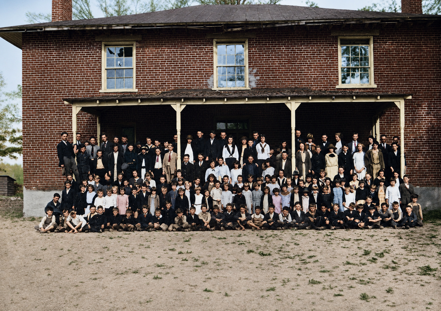 A large group of people, mostly men and boys, dressed in historical clothing, gathered on the porch and ground in front of a two-story brick building with a covered porch, in a rural outdoor setting.