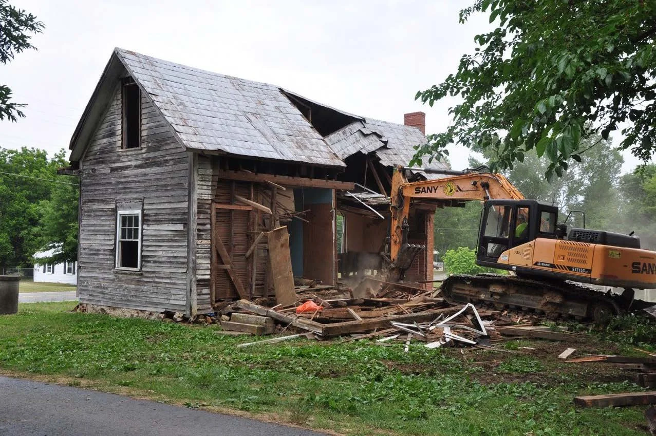 A house being demolished with an excavator tearing down the side walls, revealing the interior framework and debris.