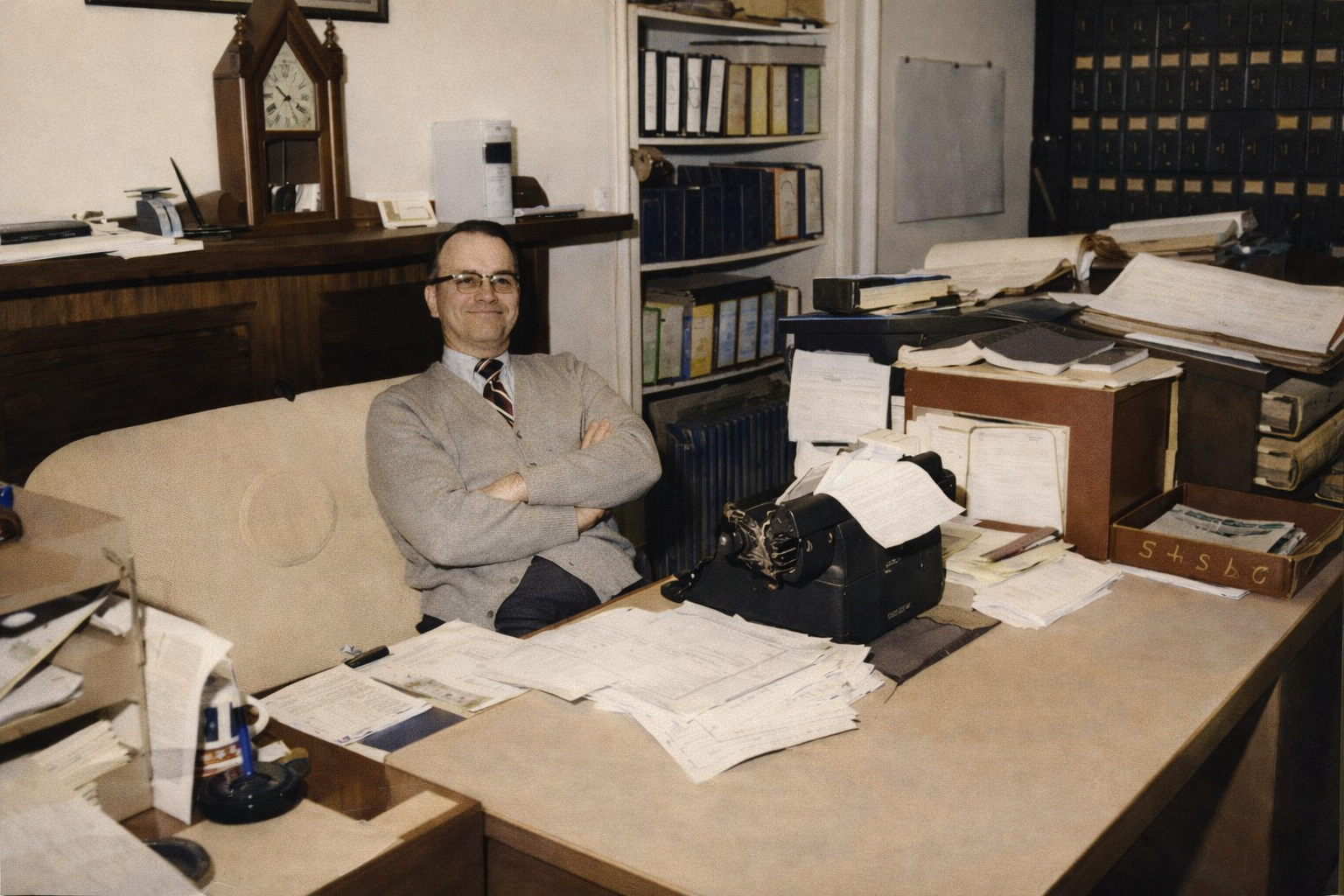 A man sitting at a cluttered office desk with arms crossed, smiling, surrounded by papers, files, and office supplies in an office with bookshelves and a clock.