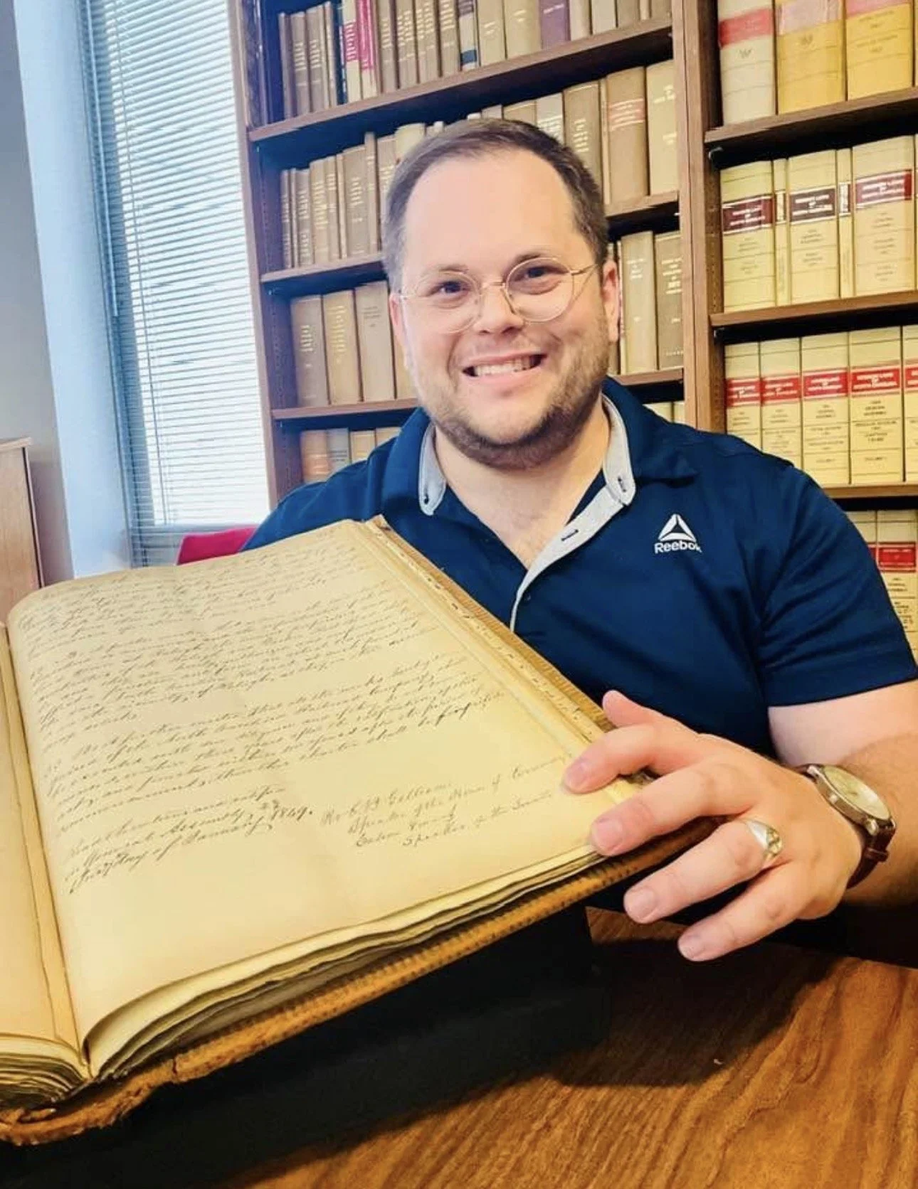 A man with glasses and a beard smiling while holding an old handwritten book open, seated at a wooden table in a library with shelves of books behind him, some with red and yellow bindings.