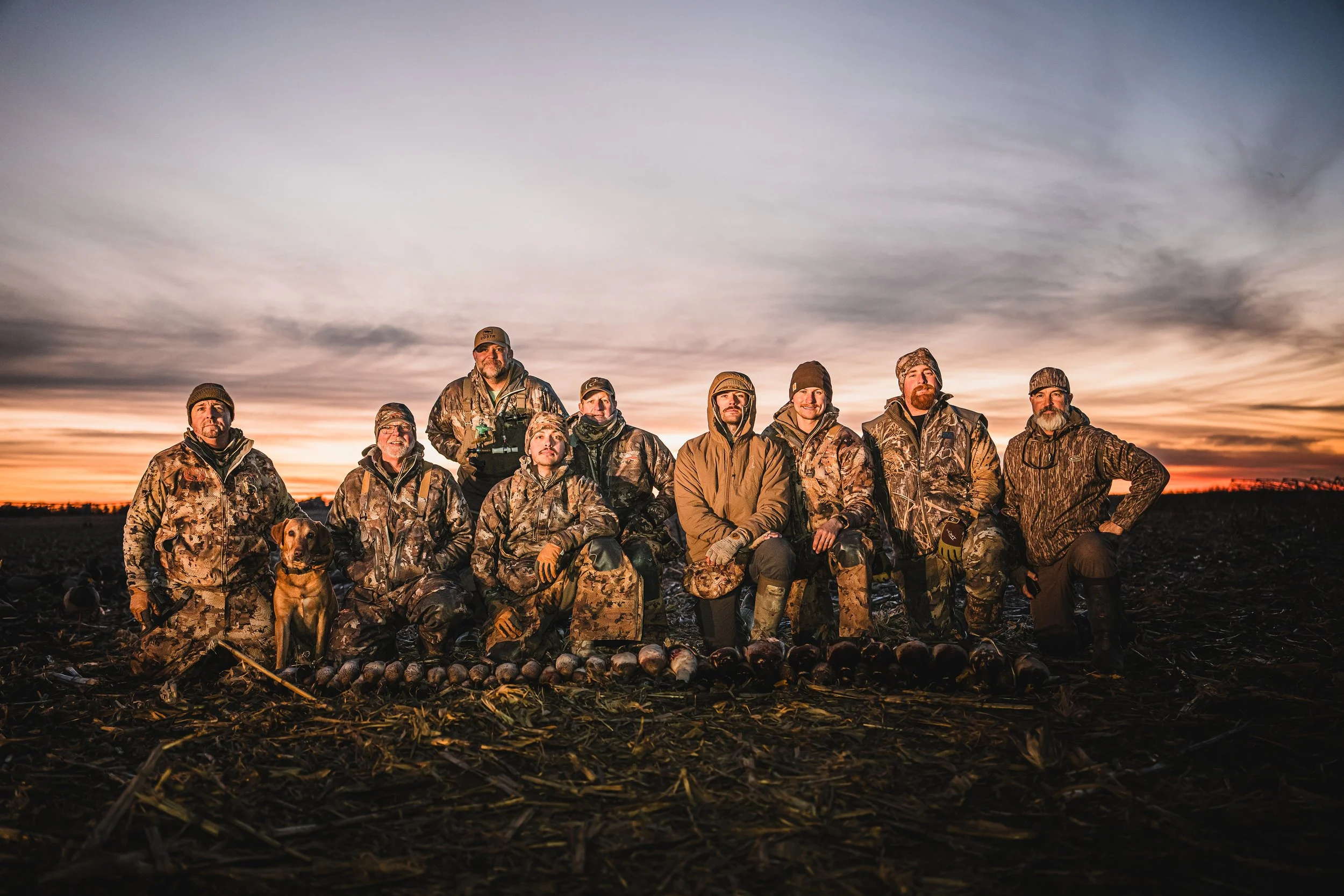 Group of ten hunters in camouflage gear posing with their hunting catch at sunrise or sunset, in a field with a dark ground and a colorful sky.