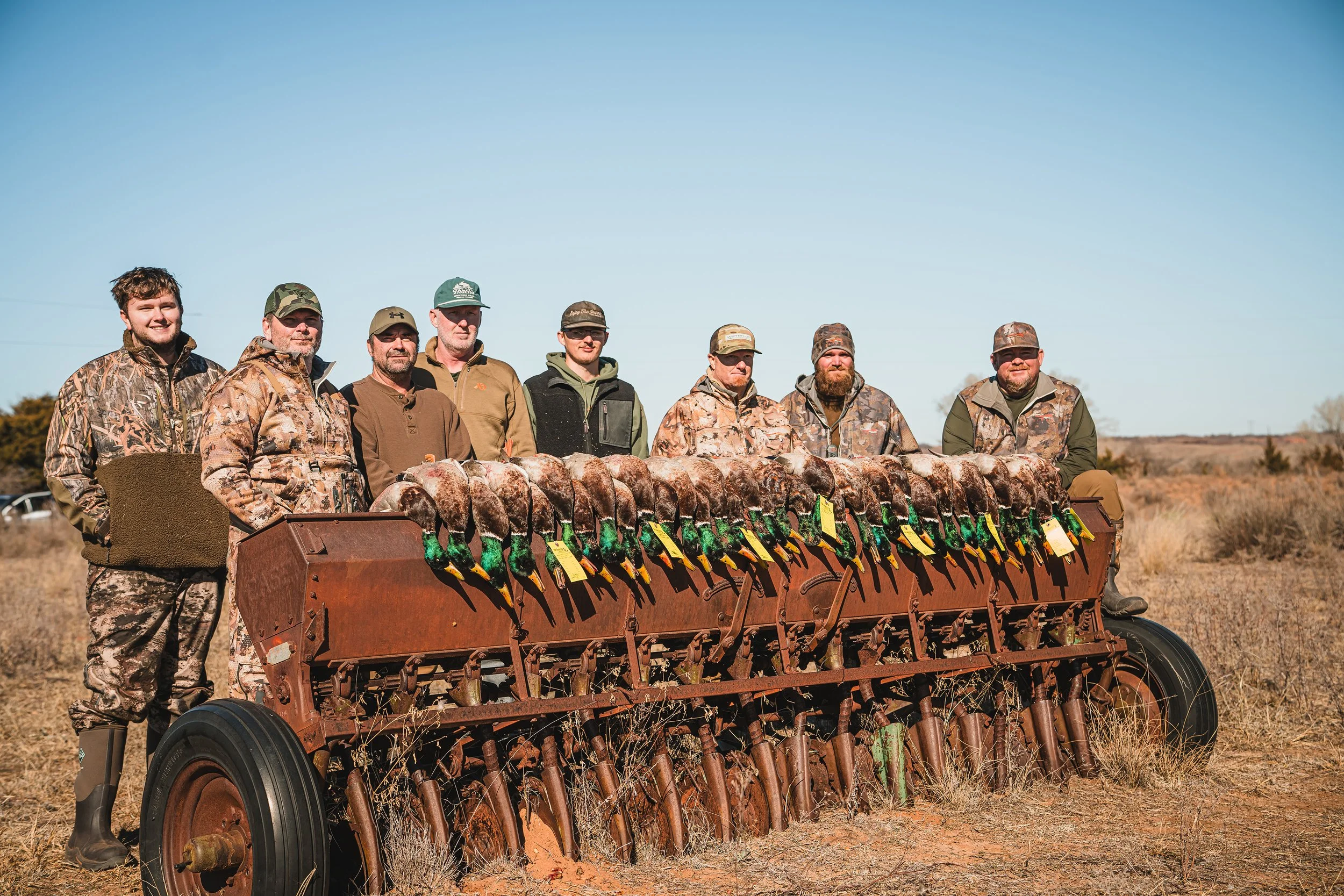 Group of nine hunters standing behind a rusty seed planter with numerous ducks attached for hunting, in an open field on a clear day.