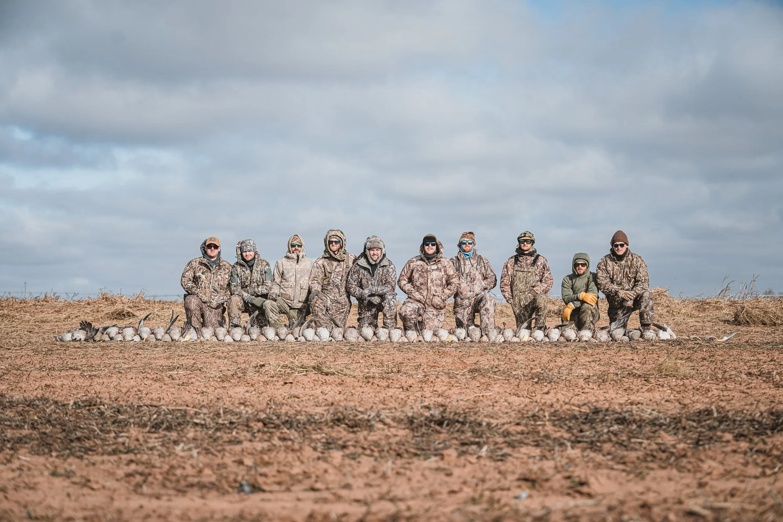 A group of ten people dressed in camouflage and outdoor gear, kneeling on the ground behind a line of deceased ducks, in an open field with cloudy sky above.