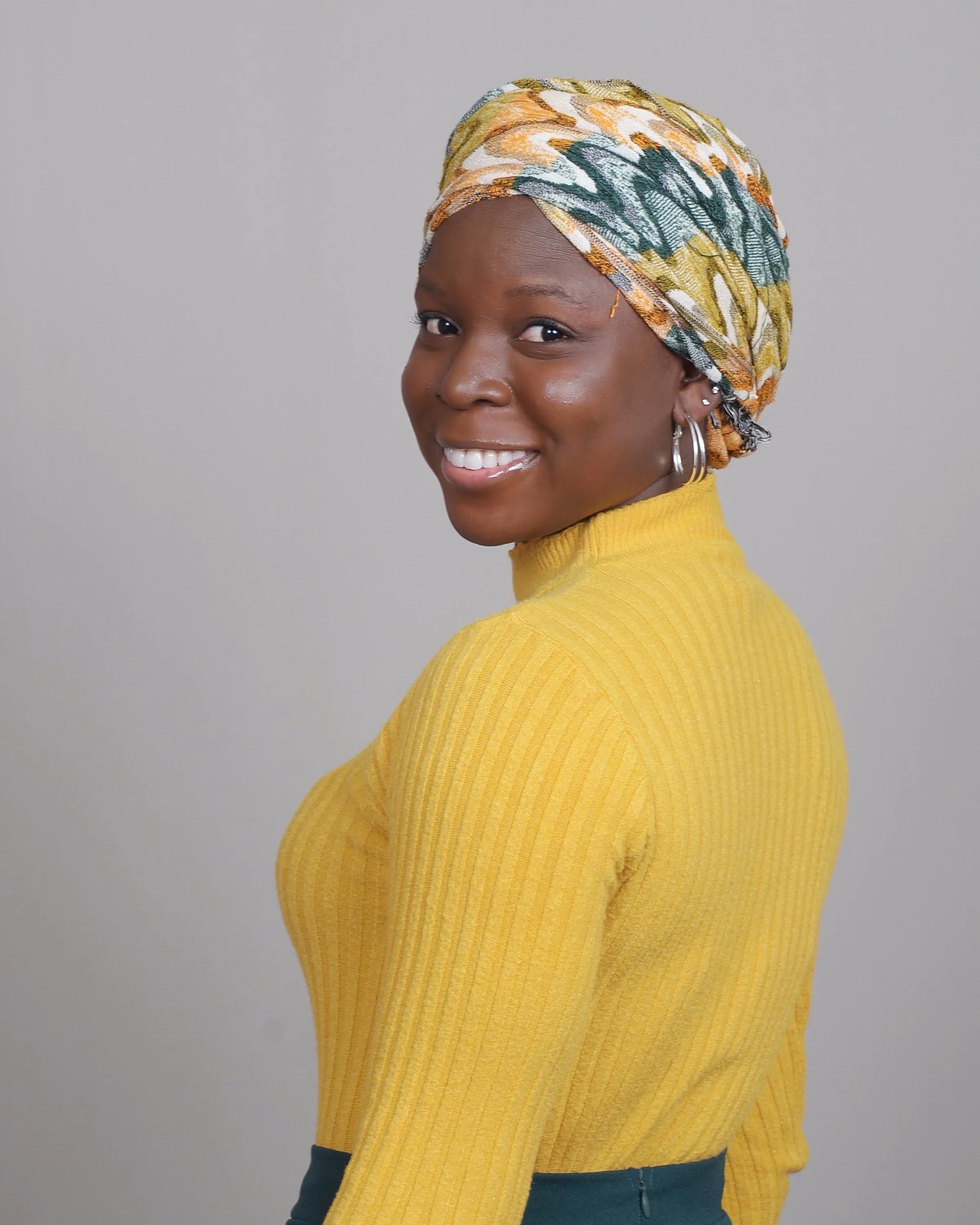 A woman smiling with a headwrap, yellow sweater, and hoop earrings, standing against a plain gray background.