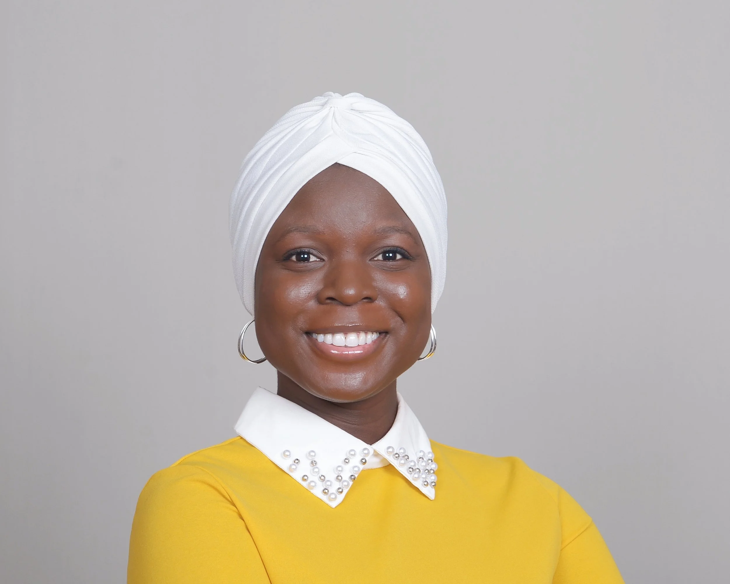 A woman with dark skin smiling, wearing a white headwrap, white collared shirt with pearl embellishments, and a yellow long-sleeve top, against a plain light gray background.