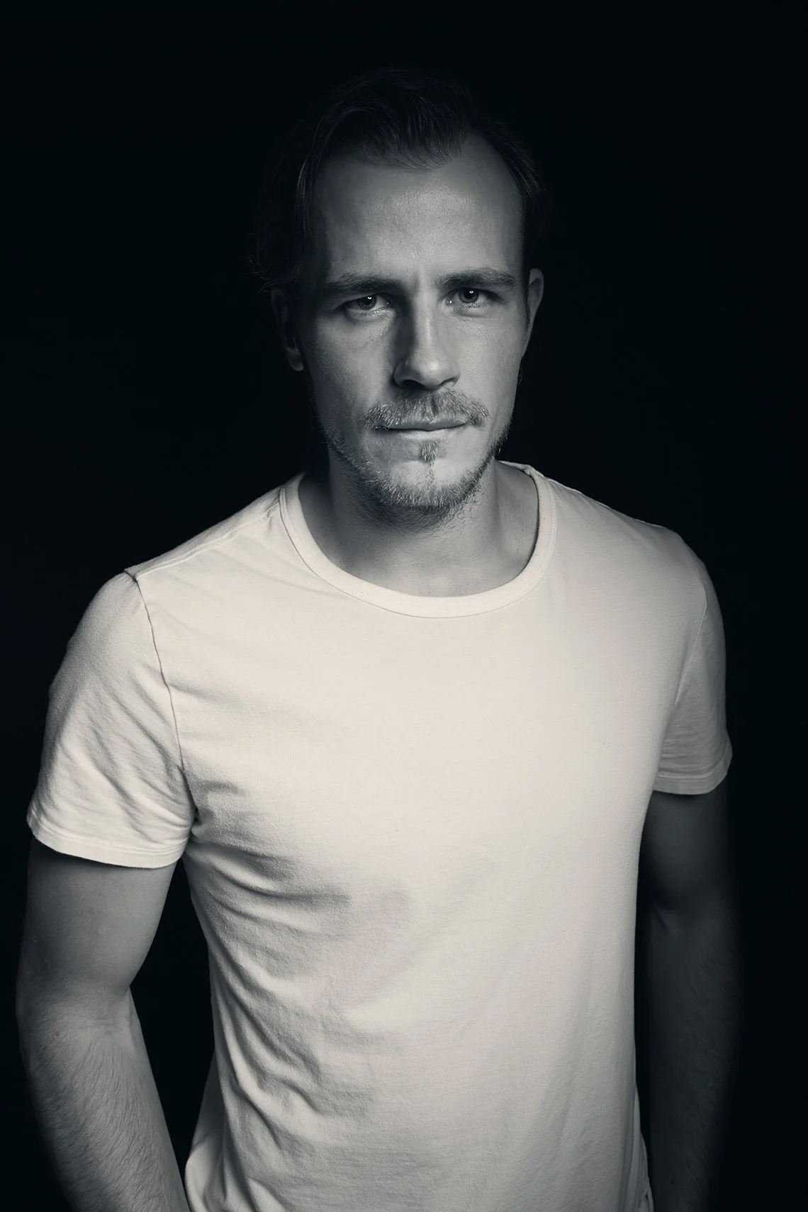 Black and white portrait of a young man with short hair and a slight beard, wearing a plain t-shirt, standing against a dark background.