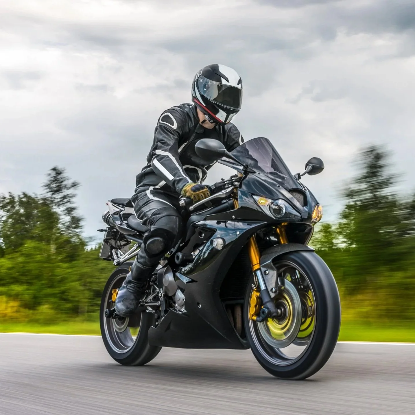 A person wearing black motorcycle gear and a helmet riding a black sport motorcycle on a road with green trees in the background on a cloudy day.