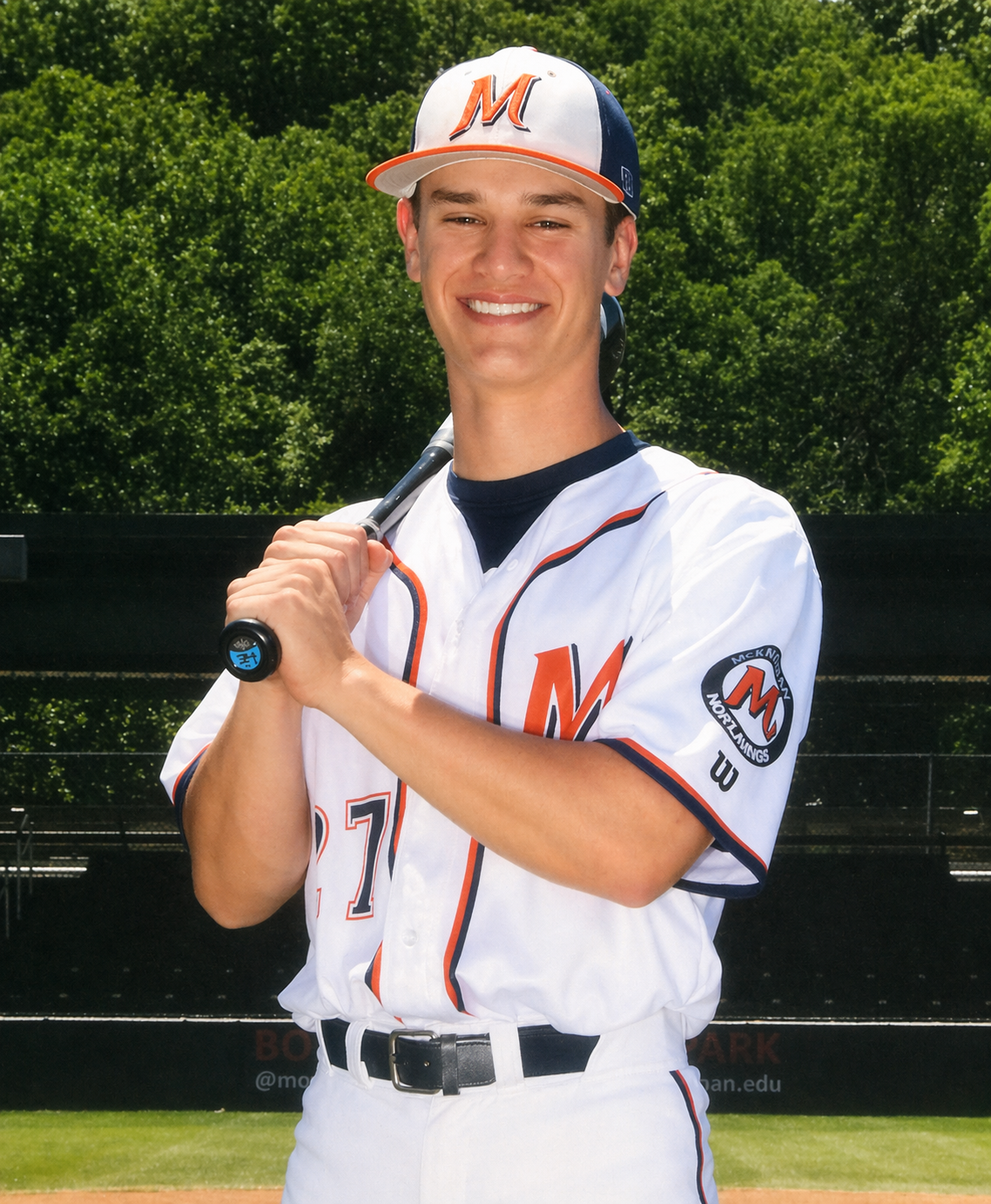 A young man in a baseball uniform holding a bat over his shoulder, smiling outdoors with trees in the background.
