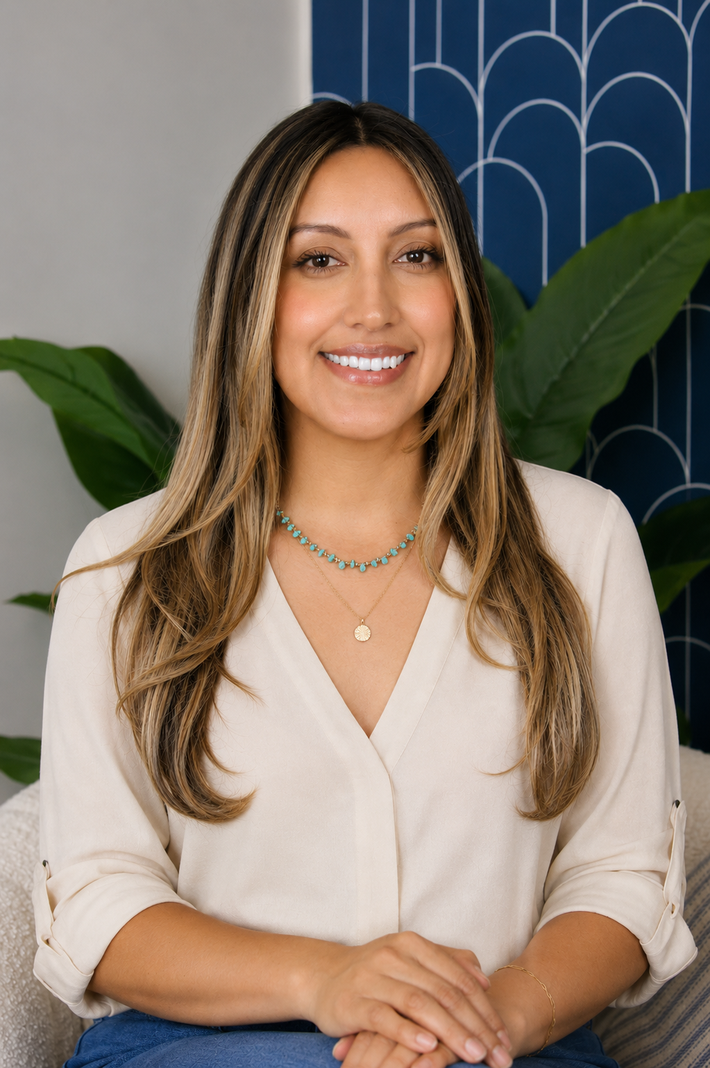 A woman with long, wavy brown hair, smiling, wearing a beige blouse, layered necklaces, and sitting indoors with green plants and a blue patterned wall in the background.