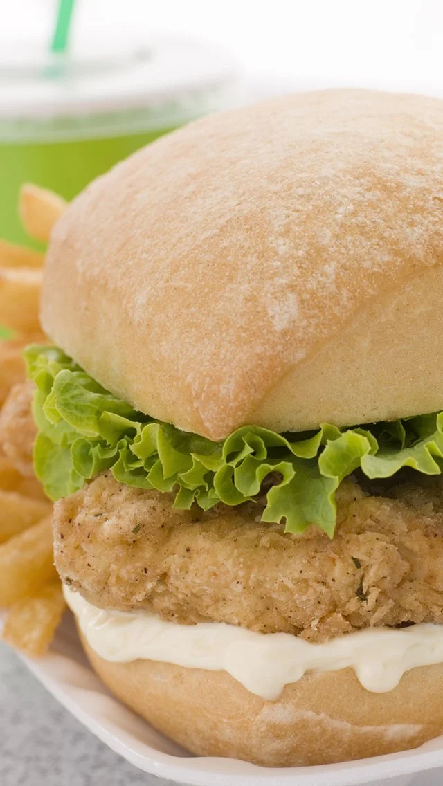 Close-up of a fried chicken sandwich with lettuce and mayonnaise in a bun, with potato chips and a green drink in the background.