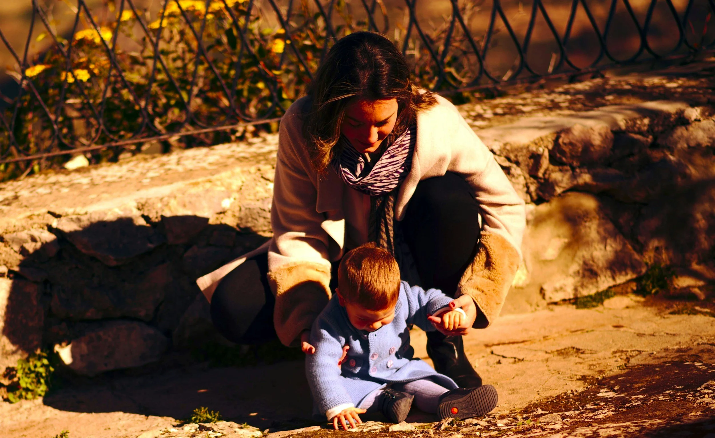A woman with dark hair, wearing a beige coat and striped scarf, crouches on the ground outside with two young children foraging or playing near a stone wall, illuminated by warm, golden sunlight.