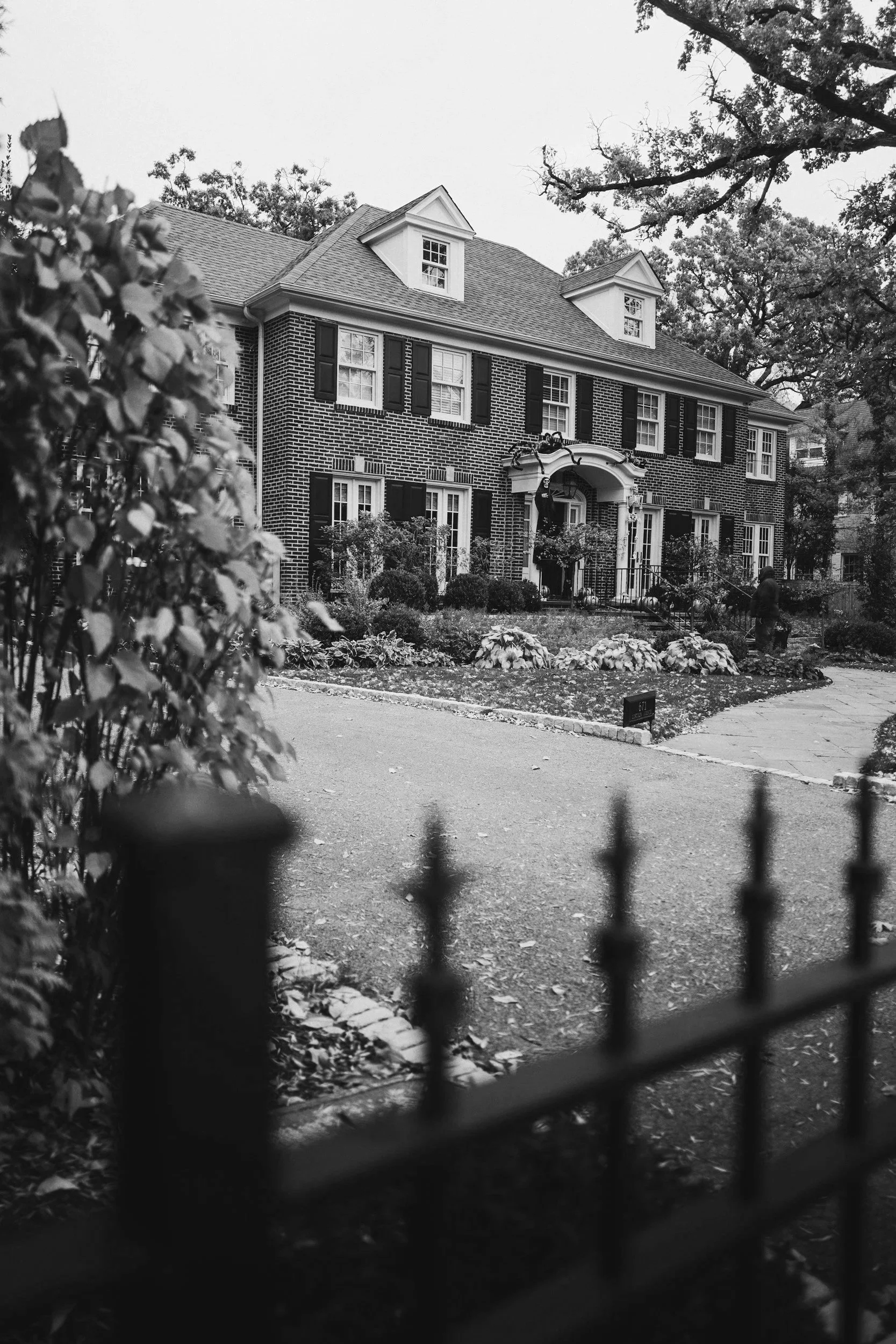 Black and white photo of a large brick house with a yard, seen through a patterned fence in the foreground. The house has multiple windows with black shutters and a small porch with decorative trim. Trees are visible in the background.