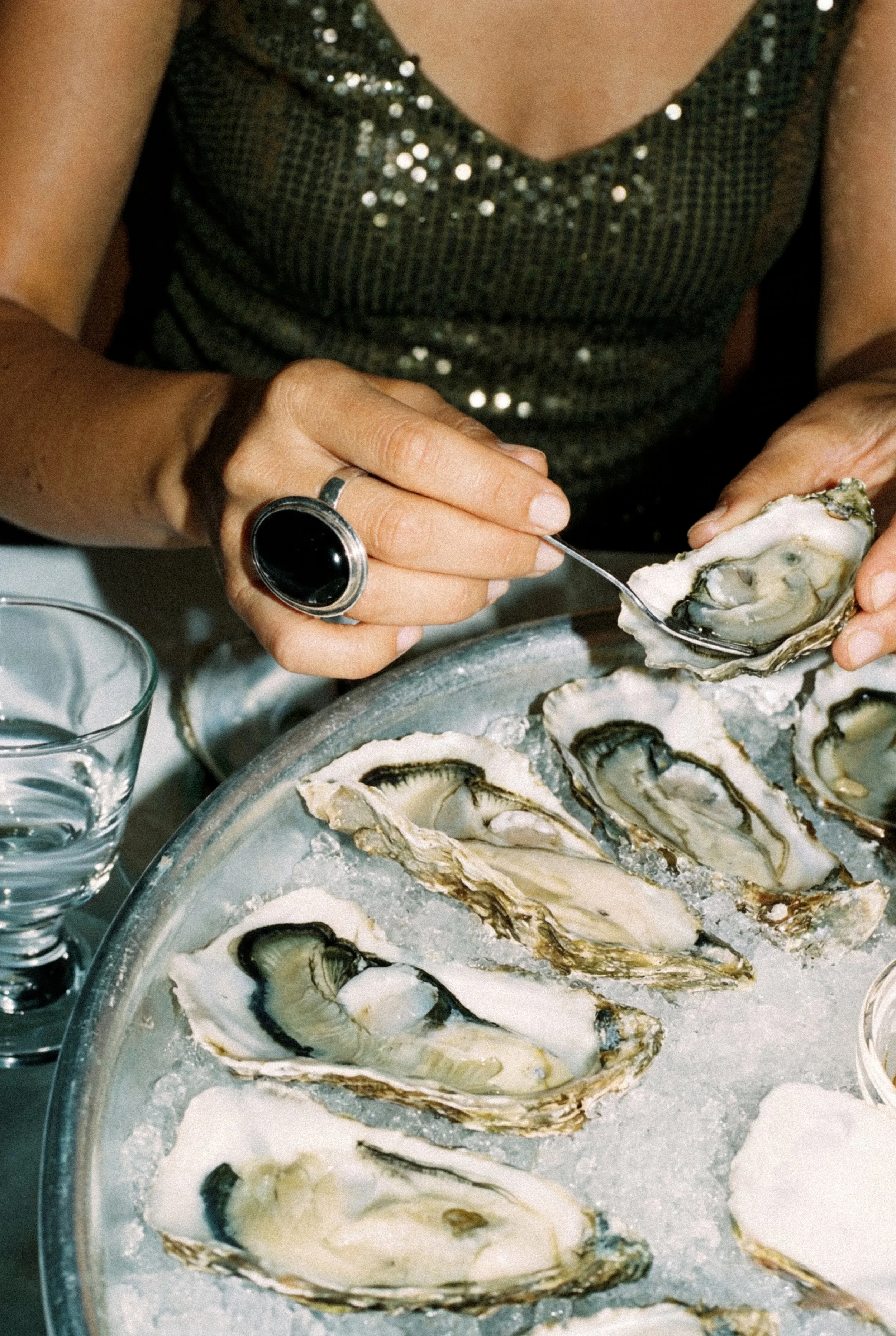 A person shucking oysters, with oysters on a tray and a glass of water nearby.