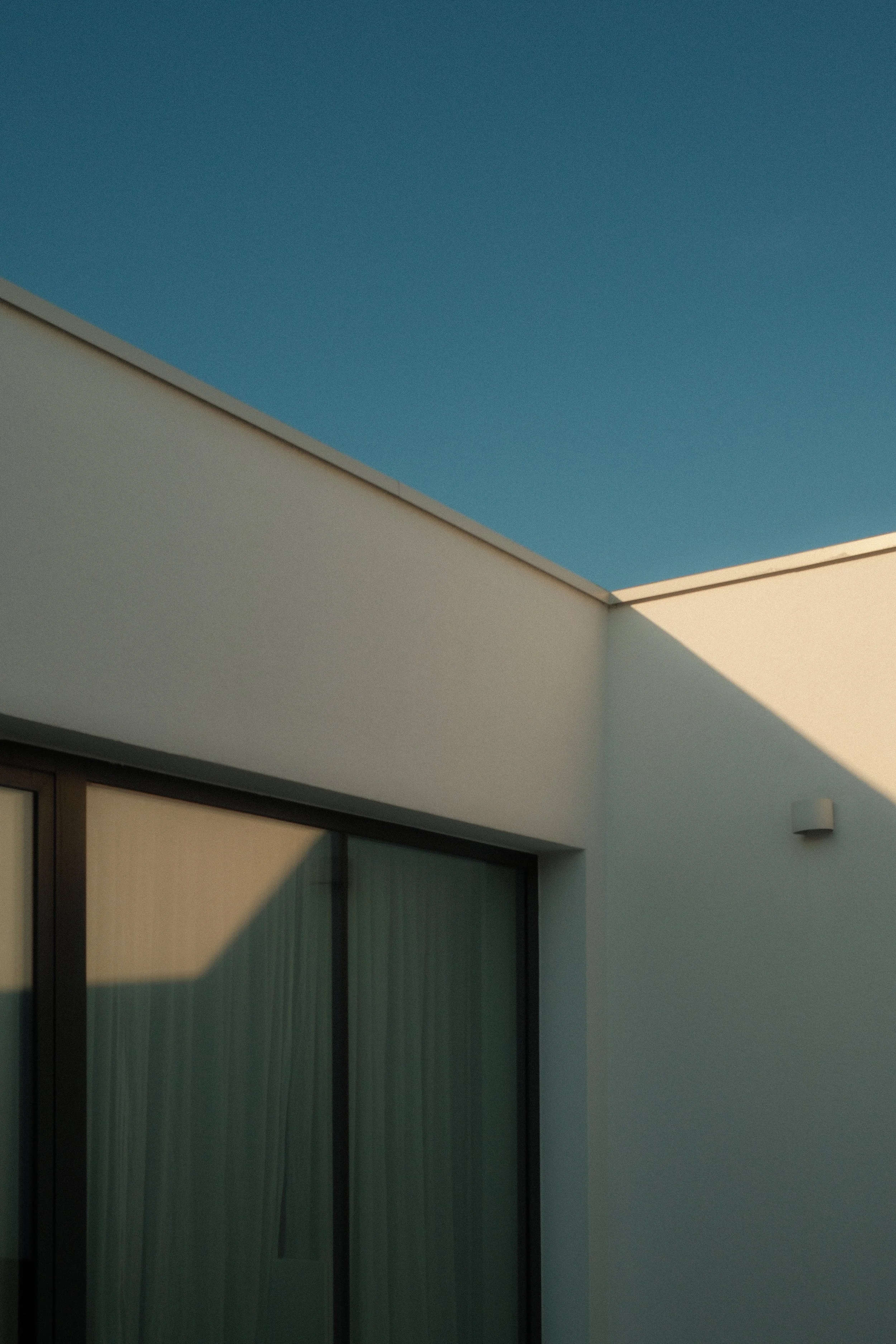 A modern building exterior with a flat roof, white walls, large glass sliding doors, and a small exterior light fixture, with a clear blue sky in the background.