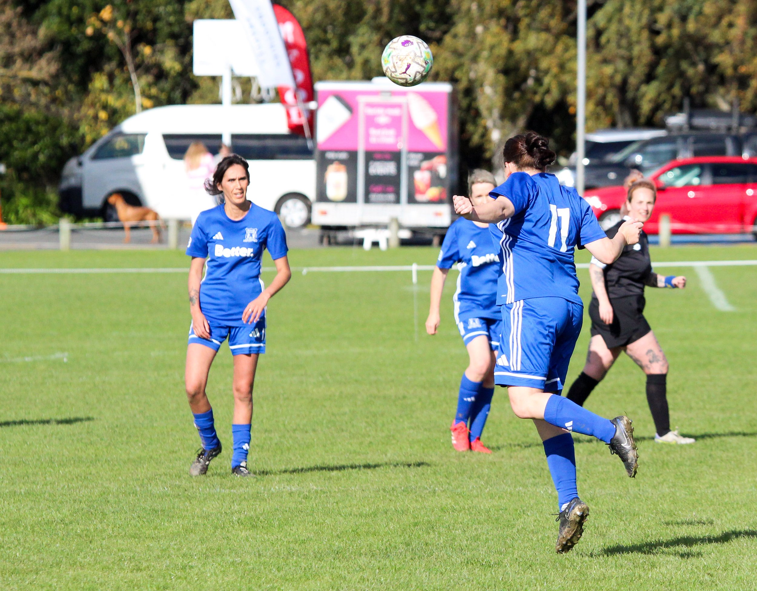 Women playing soccer on a green field, wearing blue jerseys, with a soccer ball in mid-air, and cars and trees in the background.