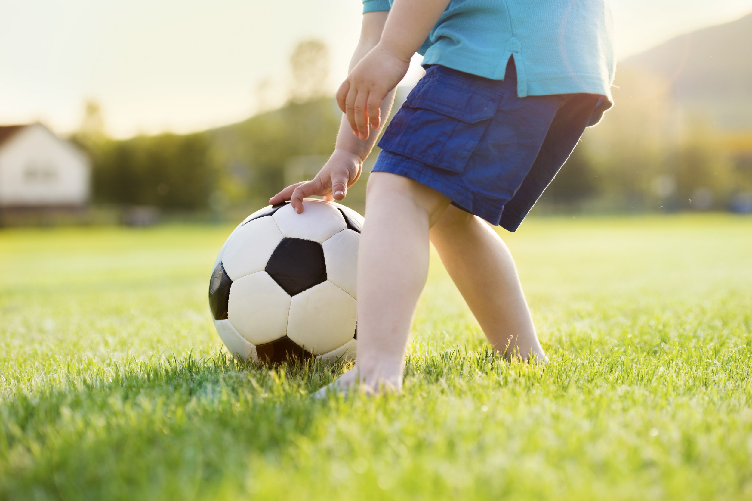 Child wearing blue shorts and a blue shirt, playing soccer on a grassy field during sunset, with a house and trees in the background.