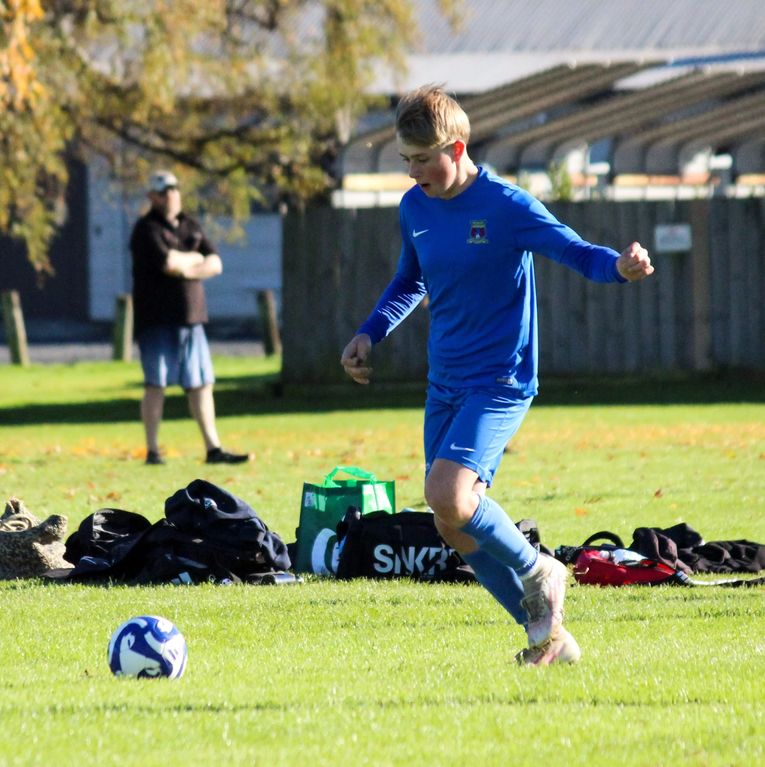Young boy in blue soccer uniform running on a grassy field with soccer balls, backpacks, and a person in the background.