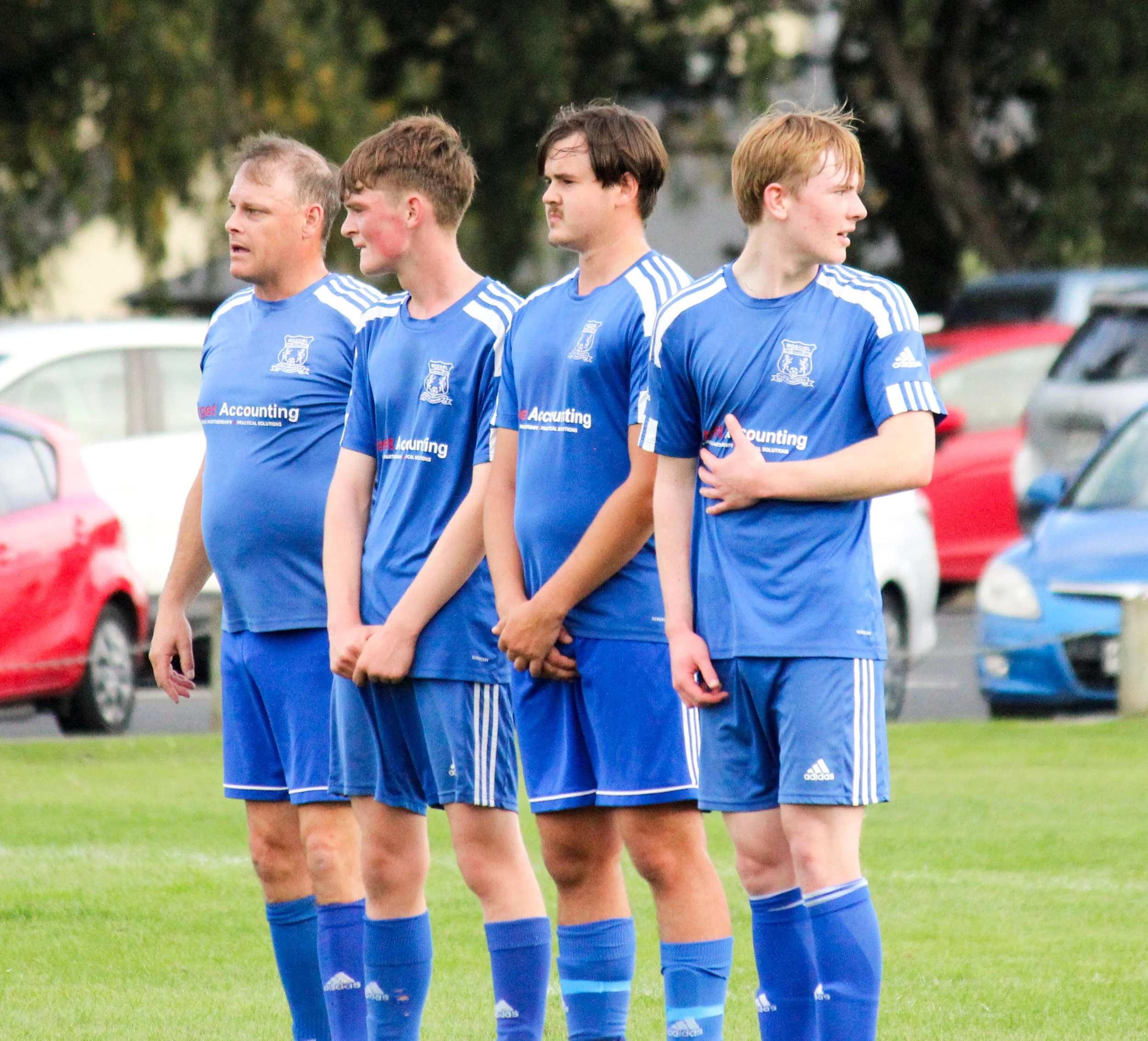 Four male soccer players standing in a row on a grassy field, wearing blue uniforms with white stripes and logos, looking to the right.