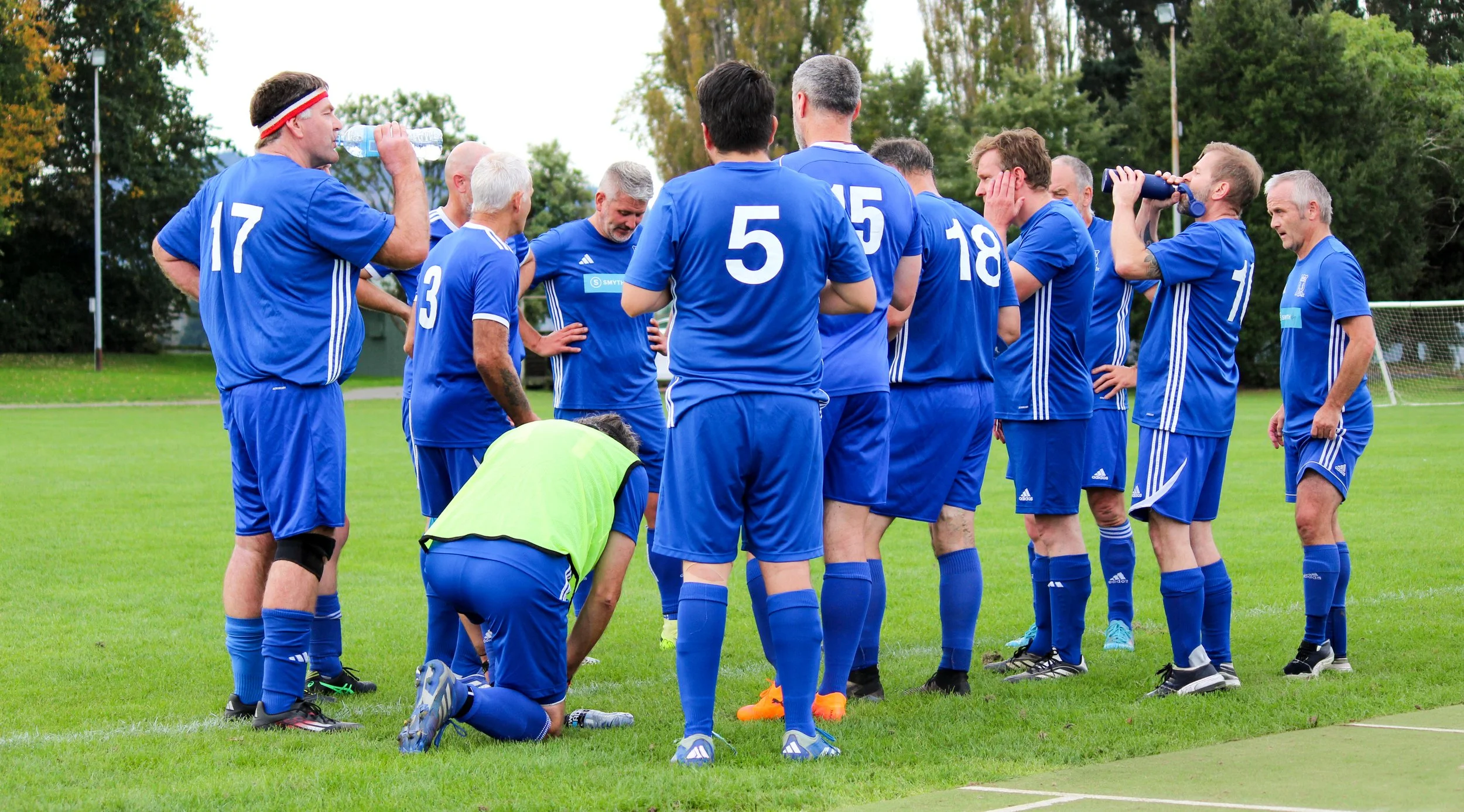 A group of soccer players in blue uniforms gathered in a huddle on the field, some drinking water, with trees in the background.